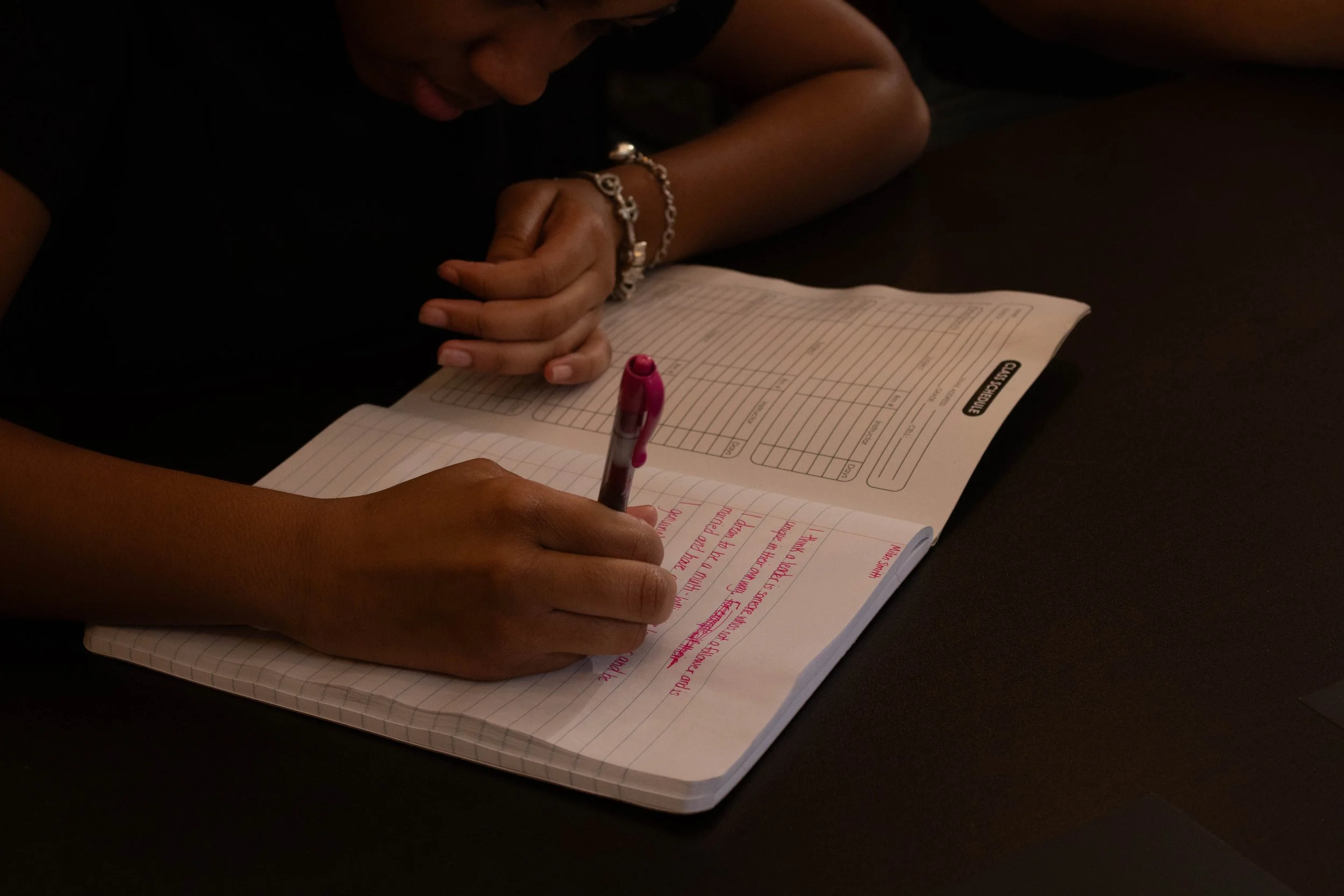 Person writing in a notebook with pink pen, with a closed book in front and another hand resting nearby.