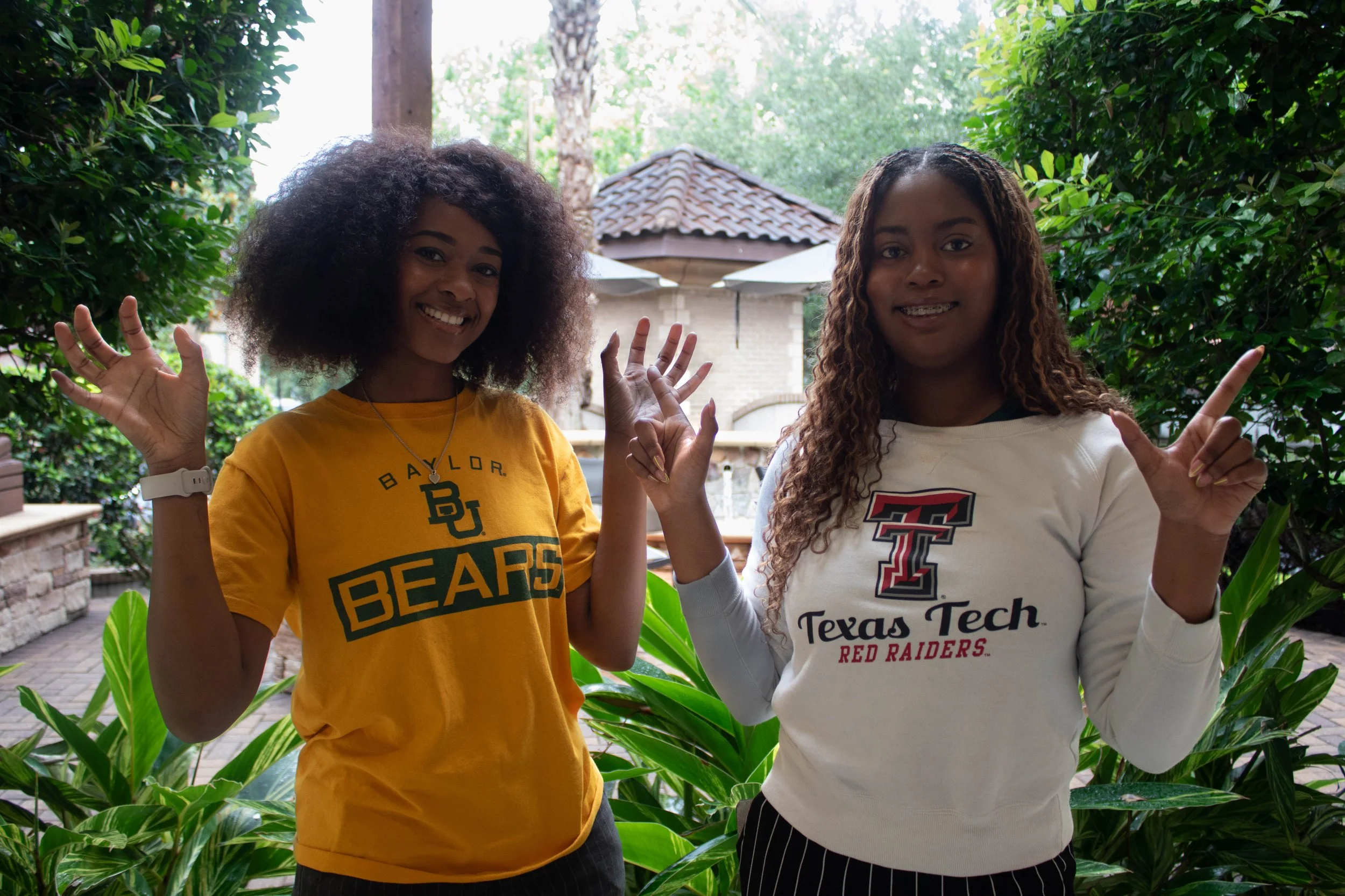 Two young women standing outdoors among green plants, smiling and waving at the camera. One is wearing a Baylor Bears T-shirt, and the other is wearing a Texas Tech Red Raiders sweatshirt.