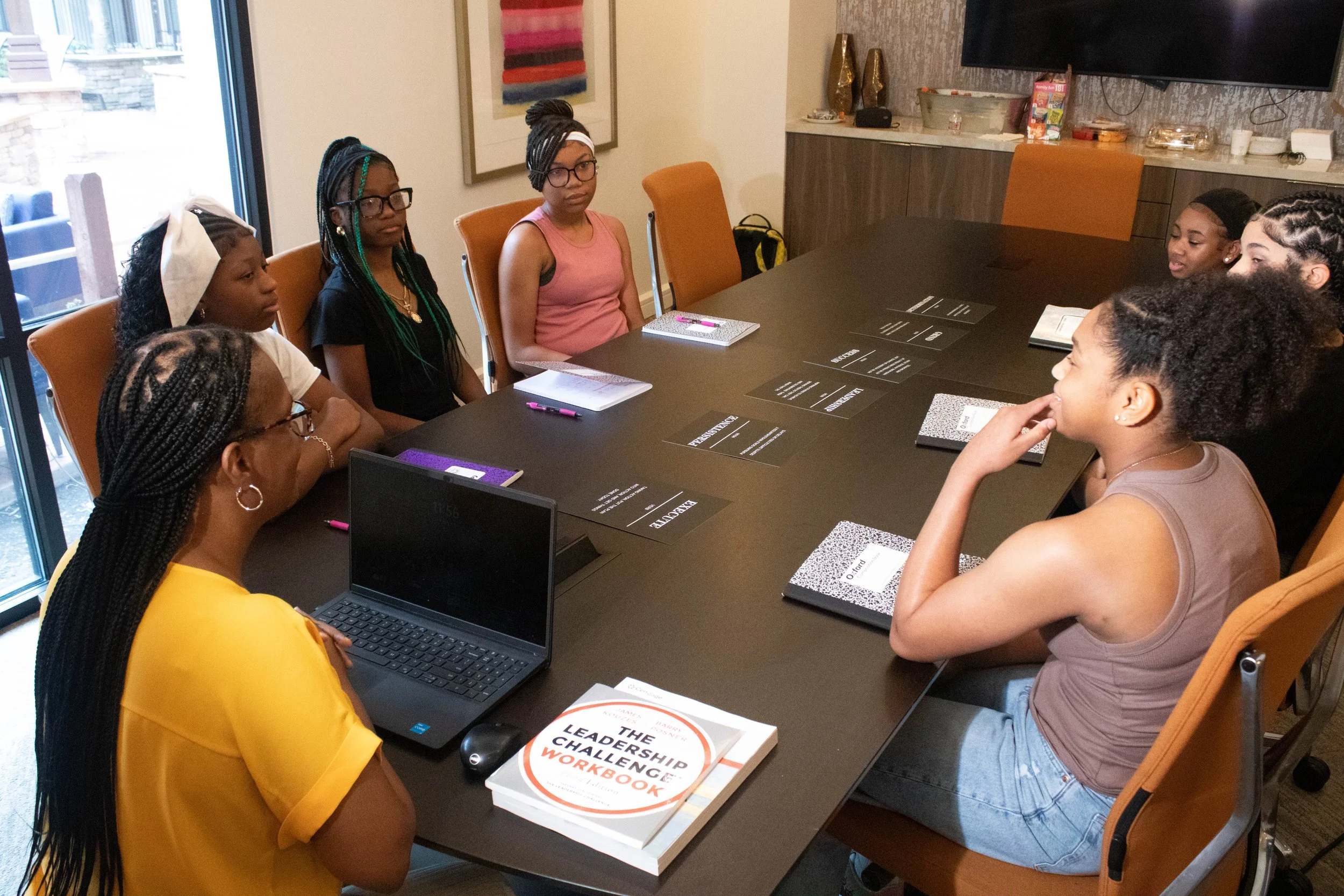 Group of women sitting around a conference table during a meeting or workshop, with notebooks, pens, laptops, and a book titled 'The Leadership Challenge' visible on the table.