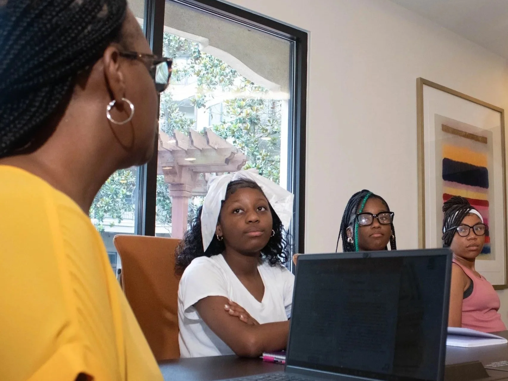 A group of women sitting in a room during a meeting or workshop; one woman in a yellow top is in the foreground, while three women, including one with a white headscarf, are in the background, with a laptop in front of them.