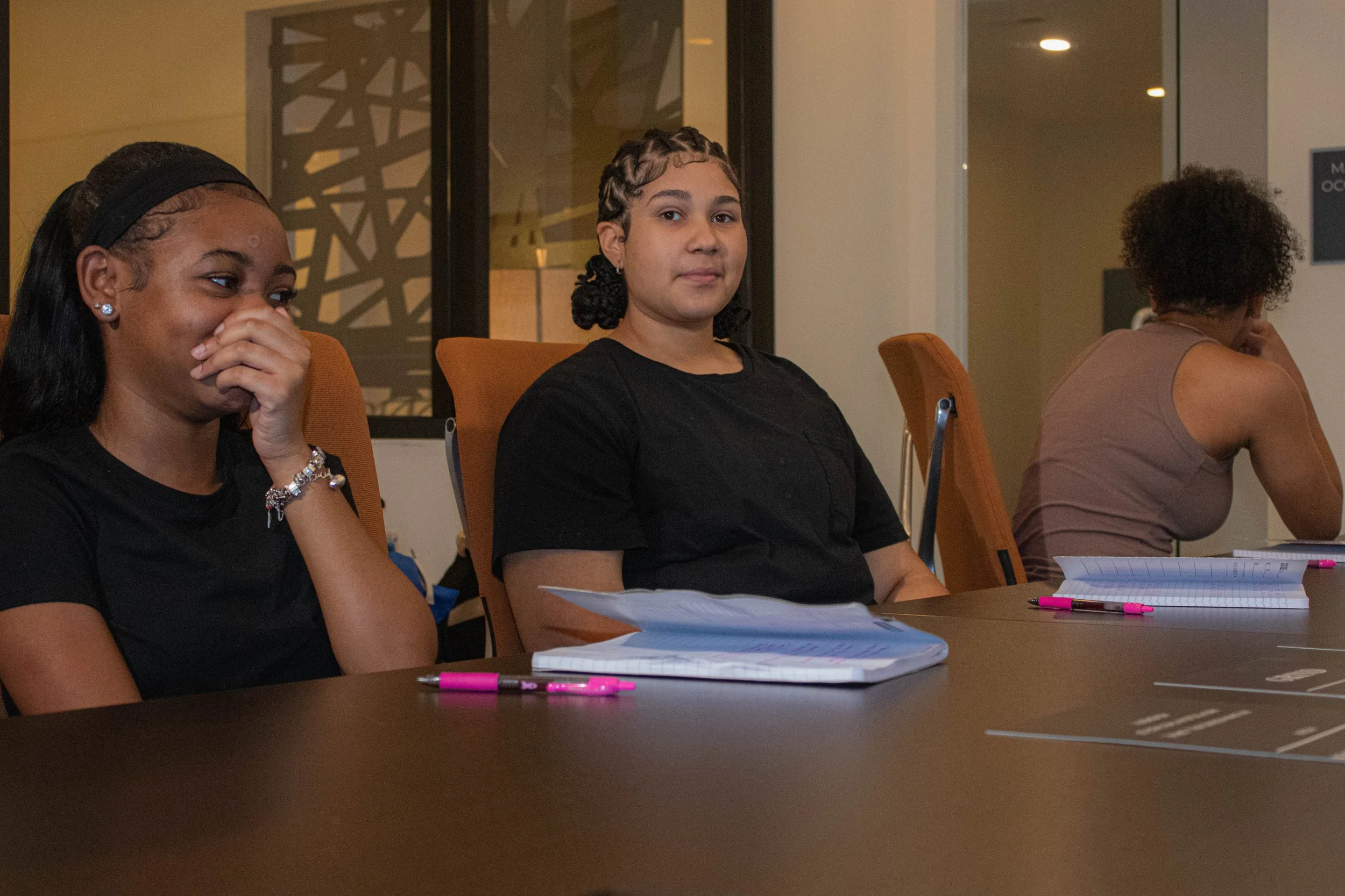 Three women sit at a conference table with notebooks and pink pens, appearing engaged in a meeting or discussion in a modern office setting.