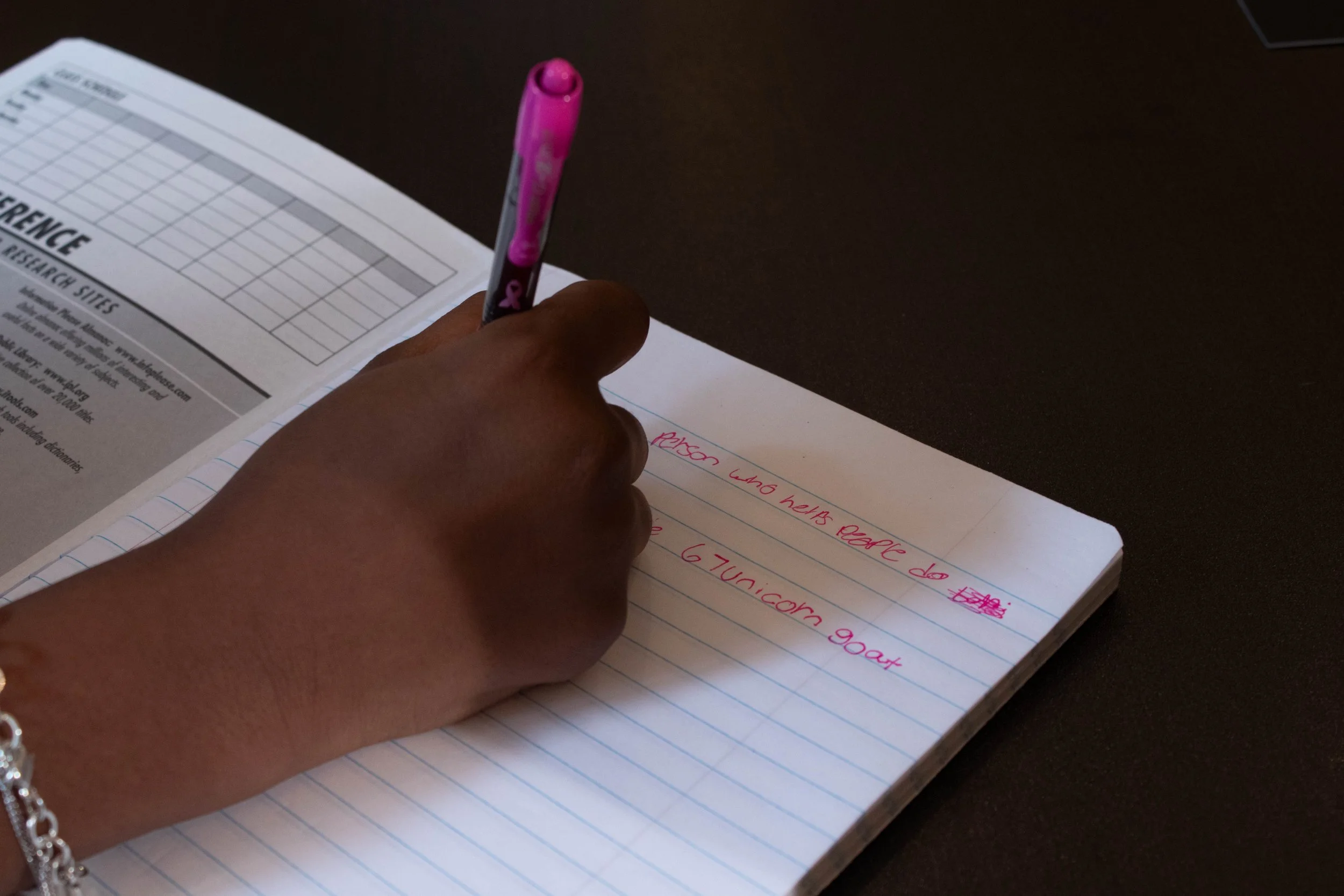 A person writing in a lined notebook with a pink marker, with an open printed research guide on the left and a dark surface around.