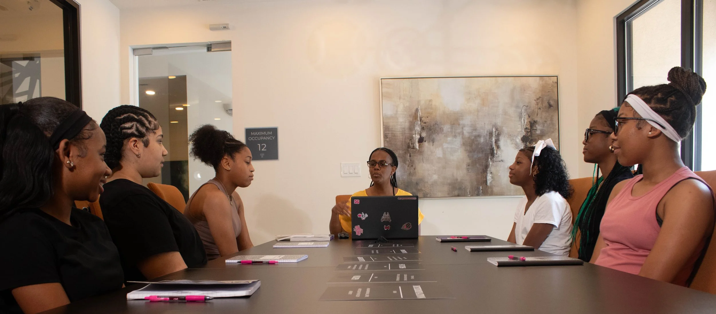 Group of women sitting around a conference table during a meeting, with notebooks and pens in front of them, in a room with large windows and abstract artwork on the wall.