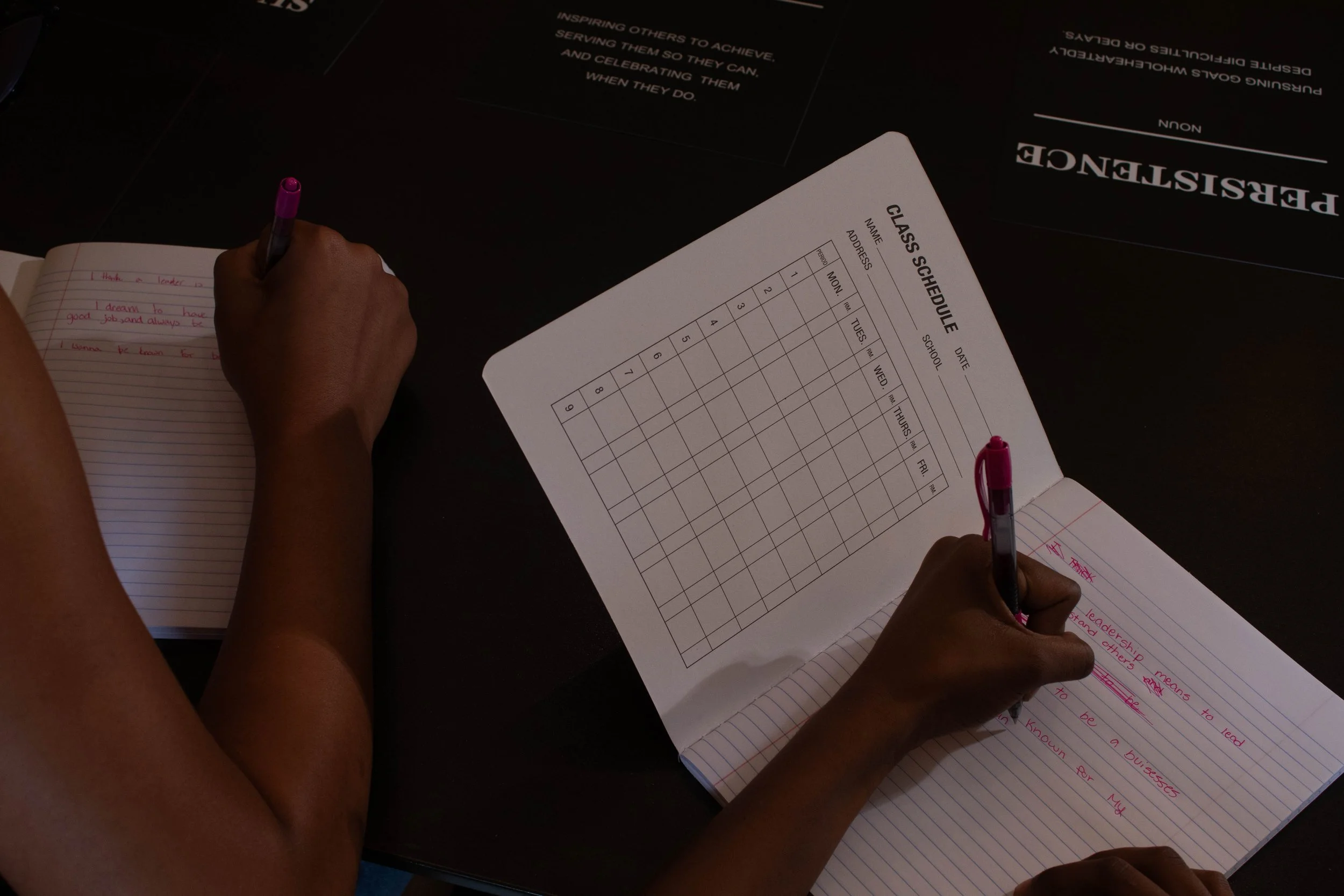 Person writing in a notebook with a pen and reviewing a class schedule on a table.