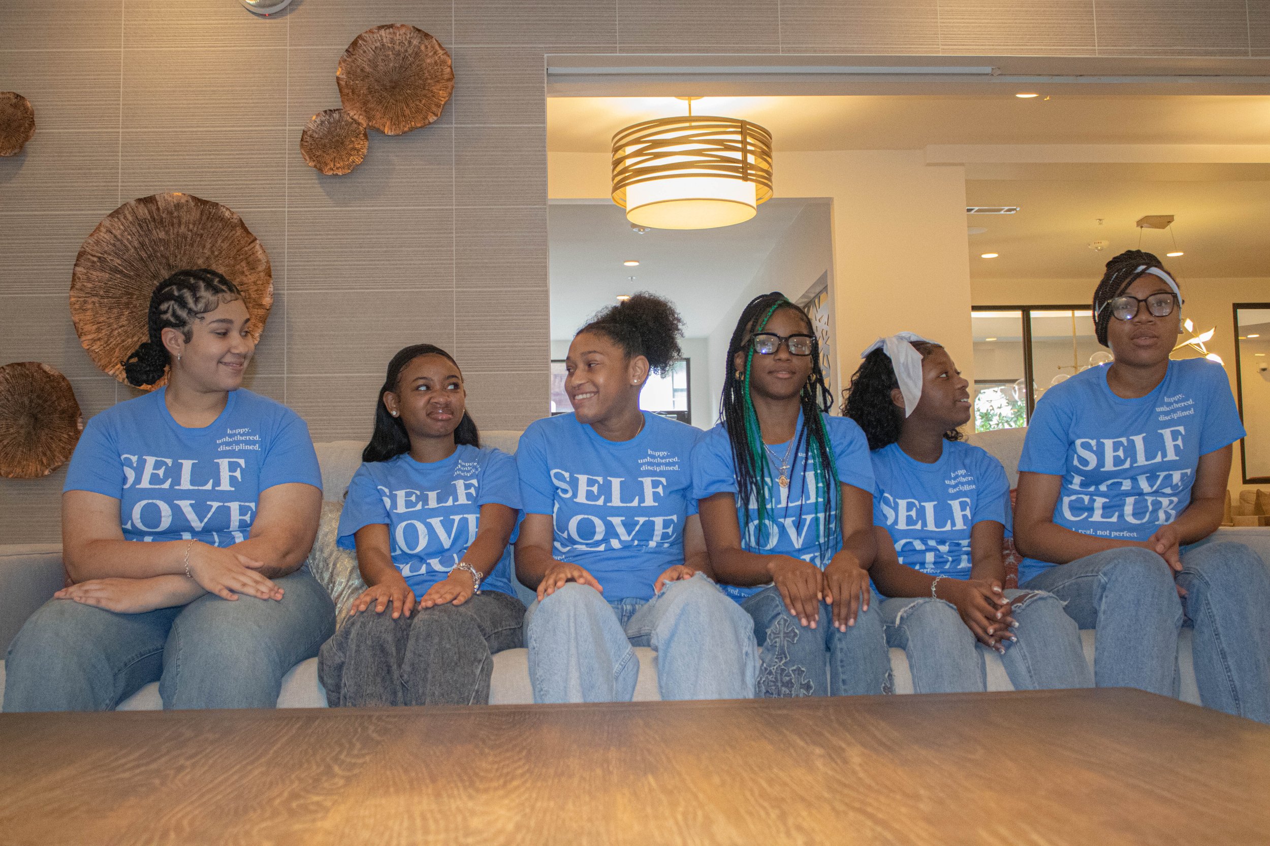 A group of six women sitting on a couch, wearing matching blue t-shirts with the words 'SELF LOVE' printed on them, in a well-lit indoor space with decorative wall art and a modern ceiling light.