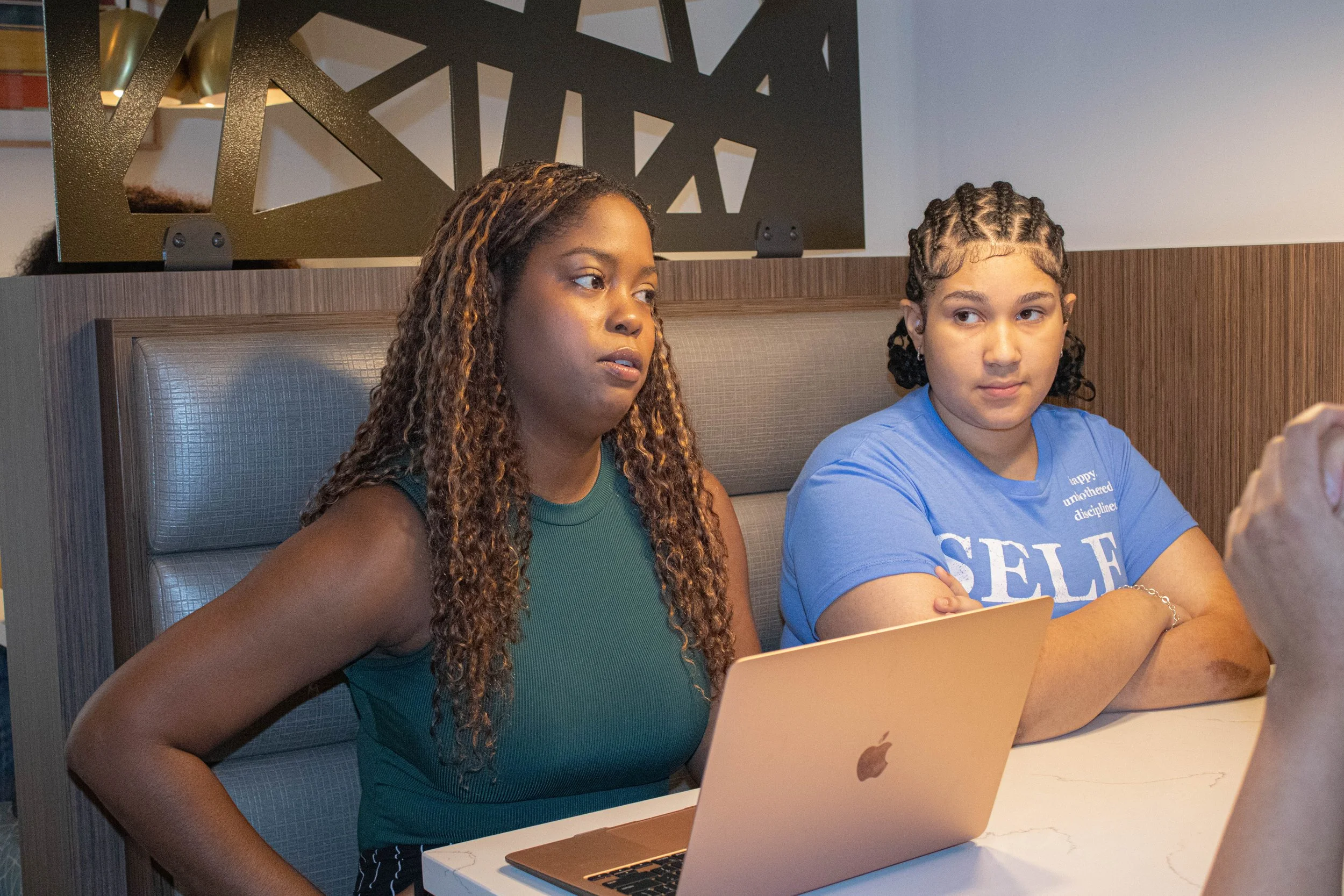Two young women sitting at a table with laptops in a modern indoor setting. The woman on the left has long, curly hair and wears a sleeveless, teal top. The woman on the right has short, braided hair and wears a blue T-shirt with white text. They are