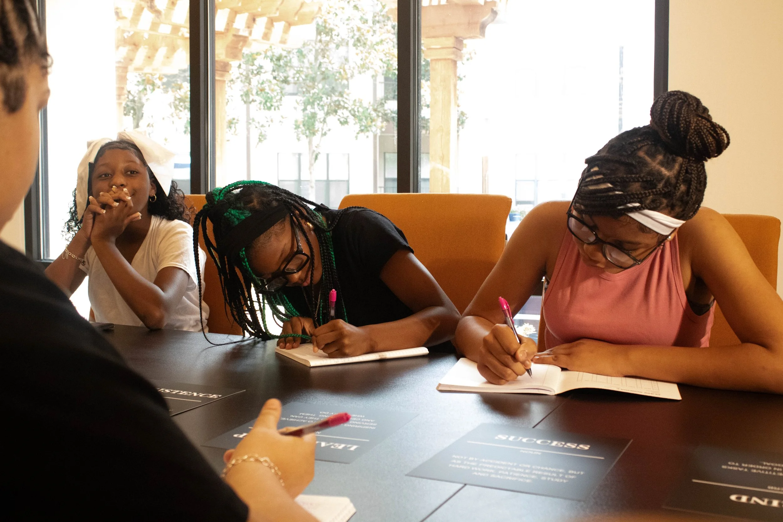 Four women sitting at a table writing in notebooks during a discussion. One woman is looking at the camera with a smile. Large windows in the background let in natural light.