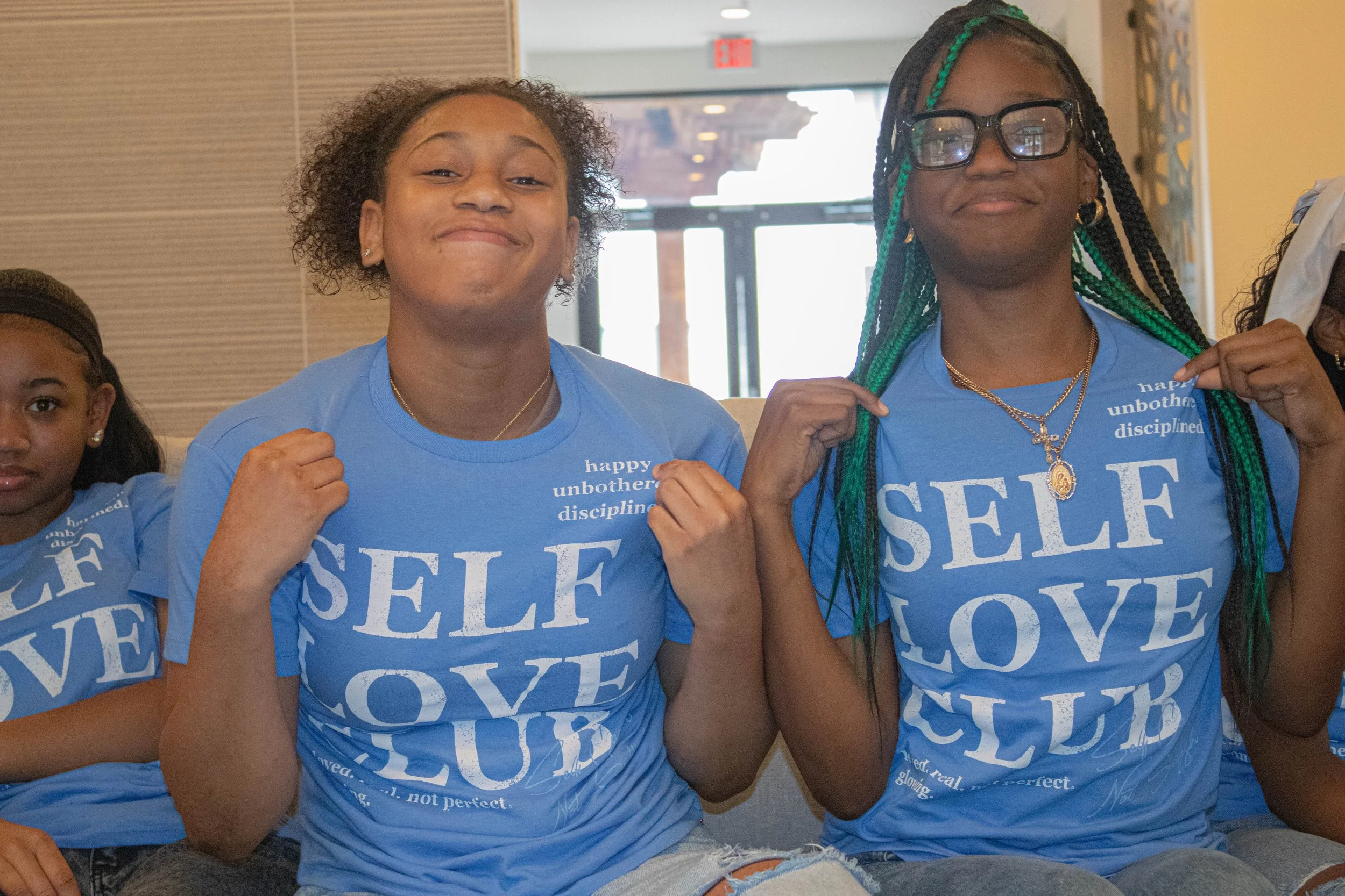 Three young girls wearing matching blue t-shirts with the words 'SELF LOVE' and other text, smiling and showing their fists in a gesture of empowerment indoors.