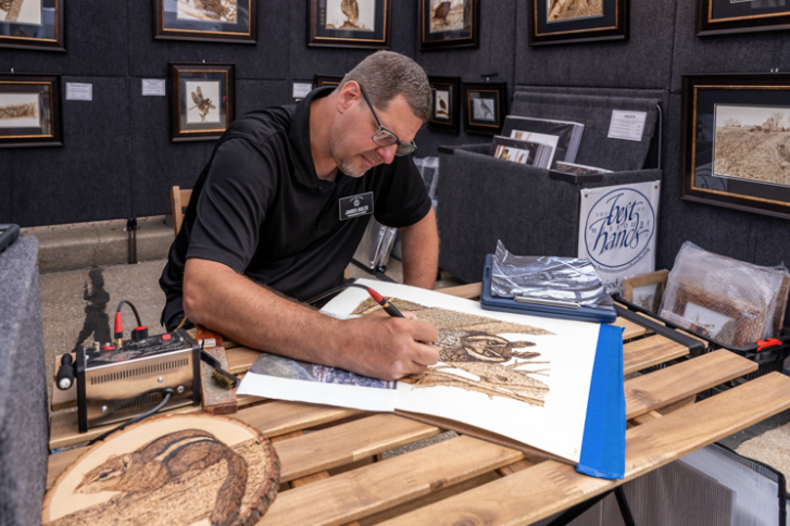 Man sitting at a wooden table, burning a nature scene with trees and wildlife, surrounded by framed artwork and supplies in an art gallery or exhibit