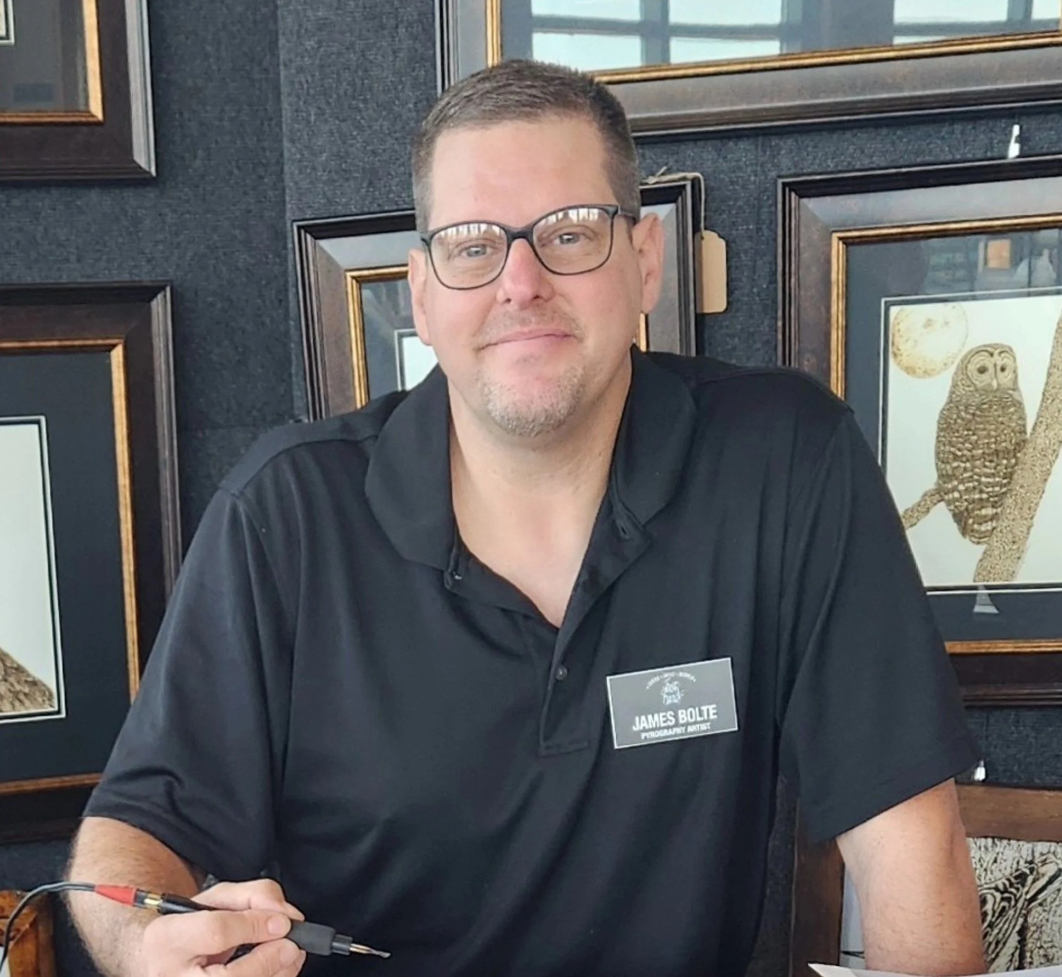 A man with glasses and short hair smiling, wearing a black shirt, standing in front of a wall with framed pictures, holding a woodburning tool in his hand, with a name tag that reads "James Bolte, Pyrography Artist."