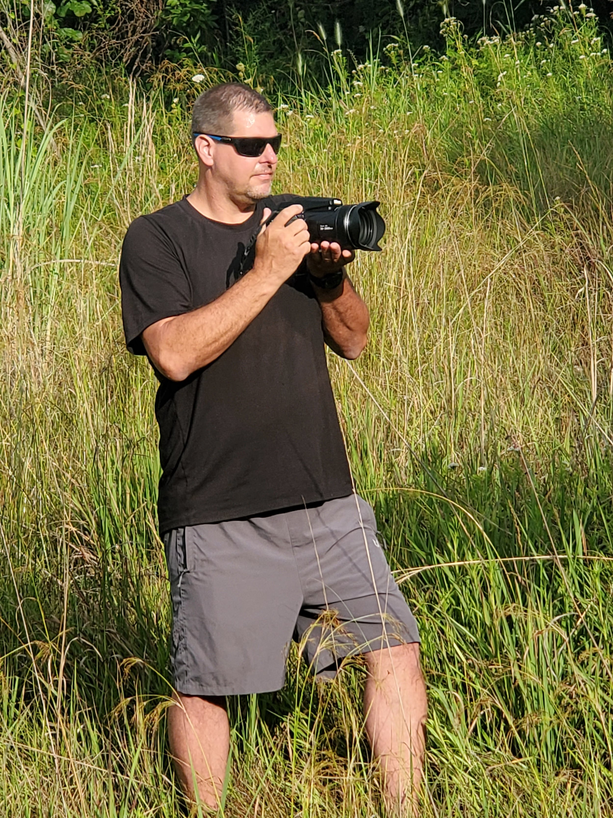 A man with short hair wearing sunglasses, a black t-shirt, and gray shorts standing in tall grass and holding a camera.