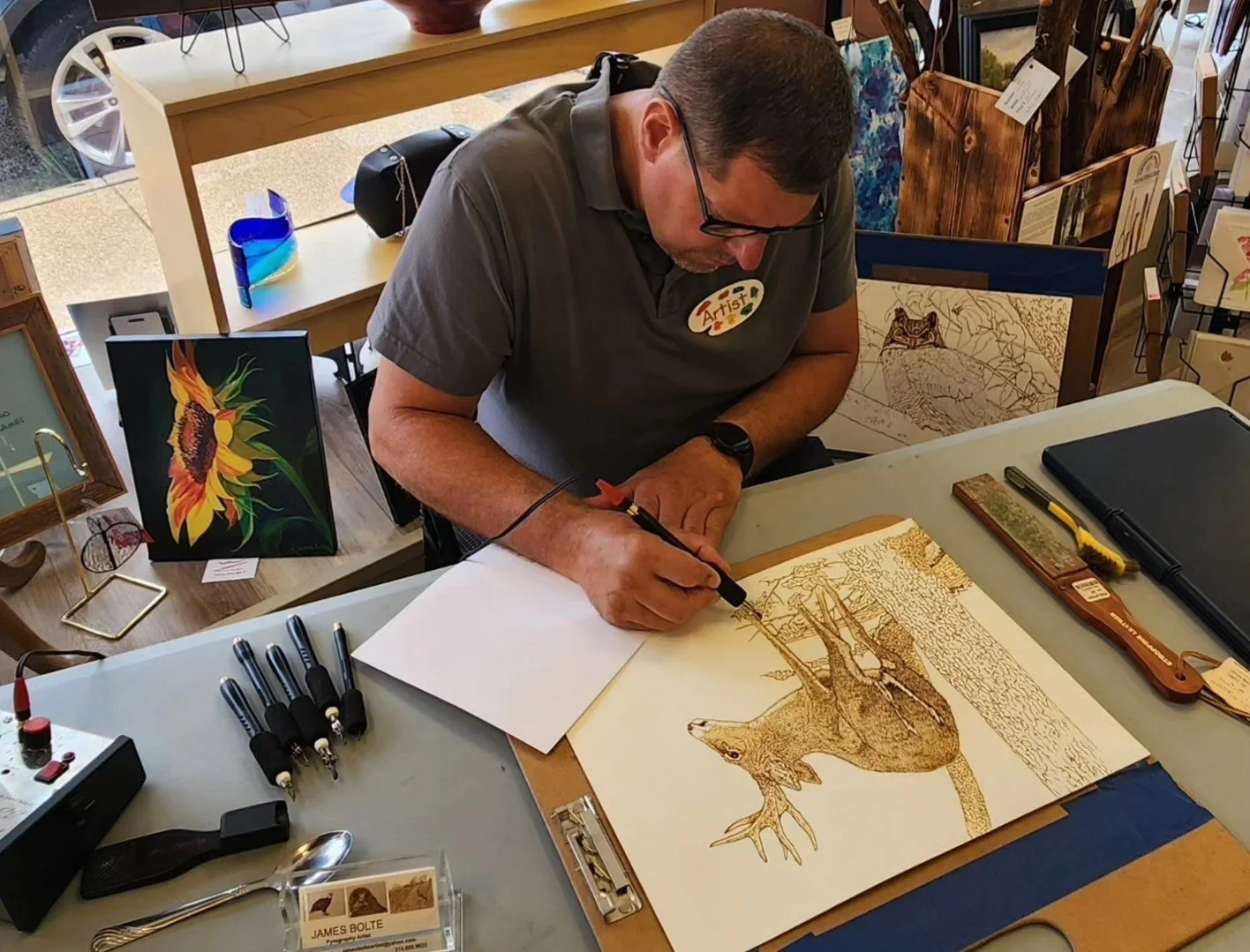An artist working on a detailed burning of a white-tailed deer on paper at a table, surrounded by art supplies and displayed artwork in the background.