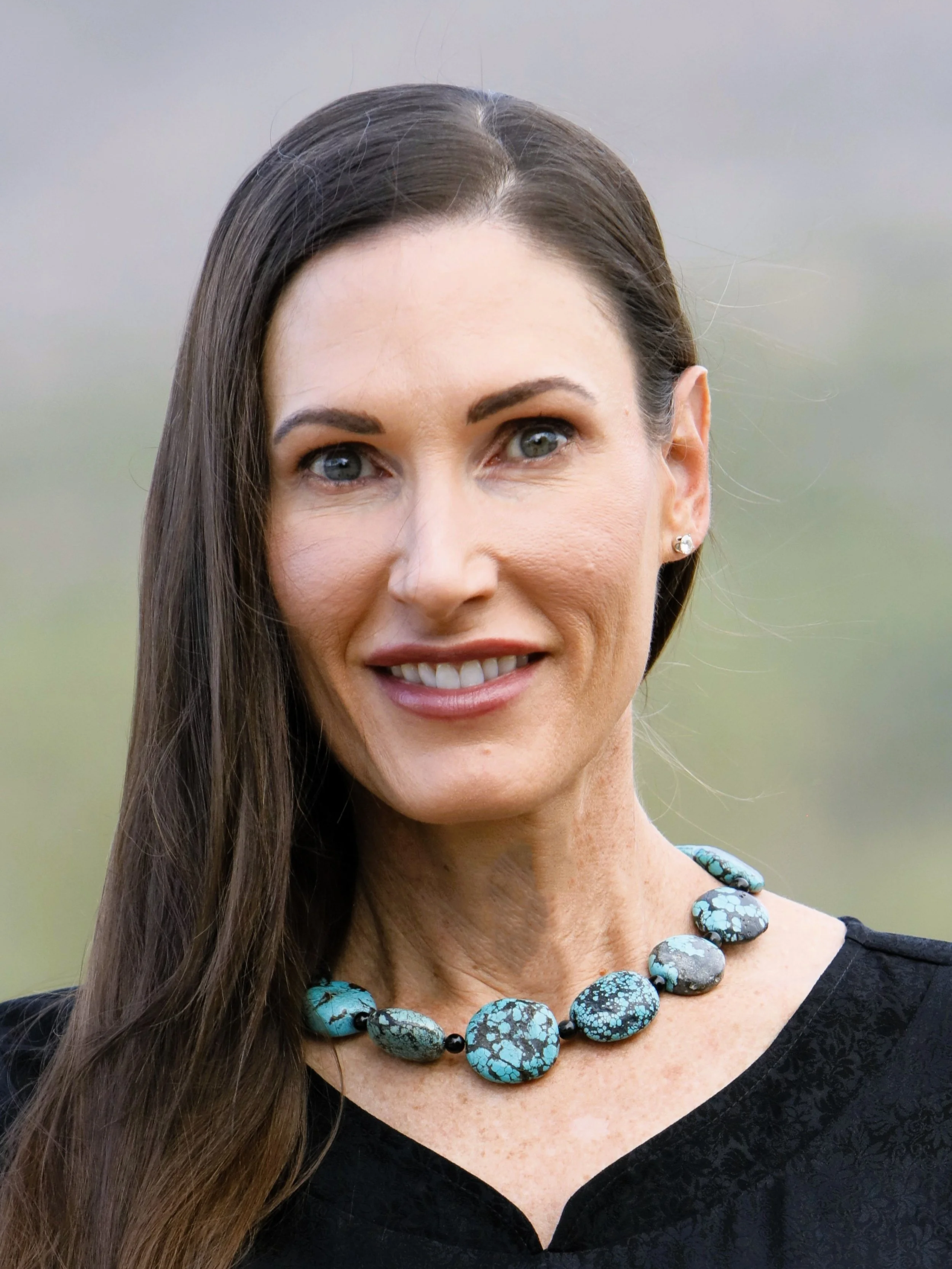 A woman with long brown hair, blue eyes, and fair skin, wearing a black top, a turquoise necklace with multiple stones, and small earrings, smiling outdoors.