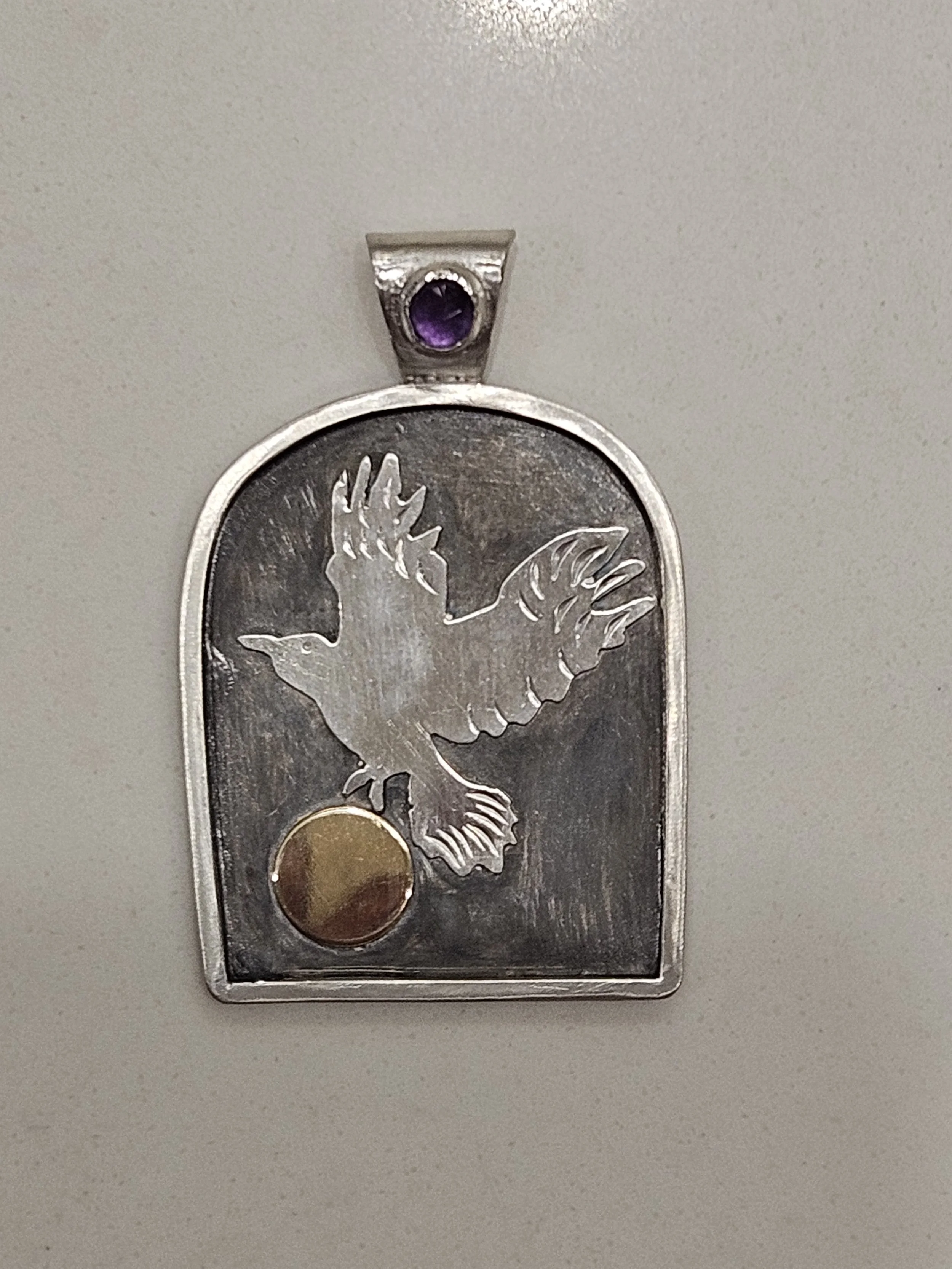 Close-up of a silver pendant with an oval amethyst gemstone at the top, a silver raven in flight and a round gold disc at the bottom, against a plain background.