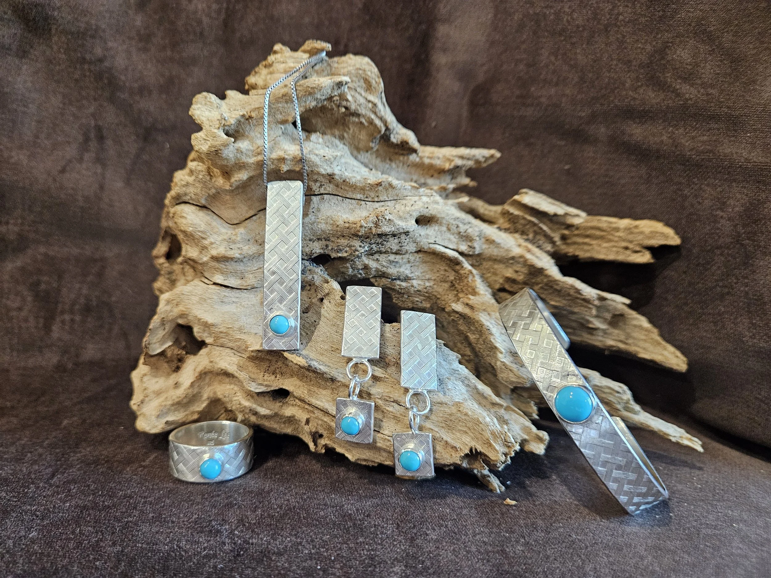Sterling silver basket weave jewelry including a ring, a bracelet, pendant and earrings all with Sleeping Beauty turquoise. Displayed against a piece of driftwood and a dark fabric background.