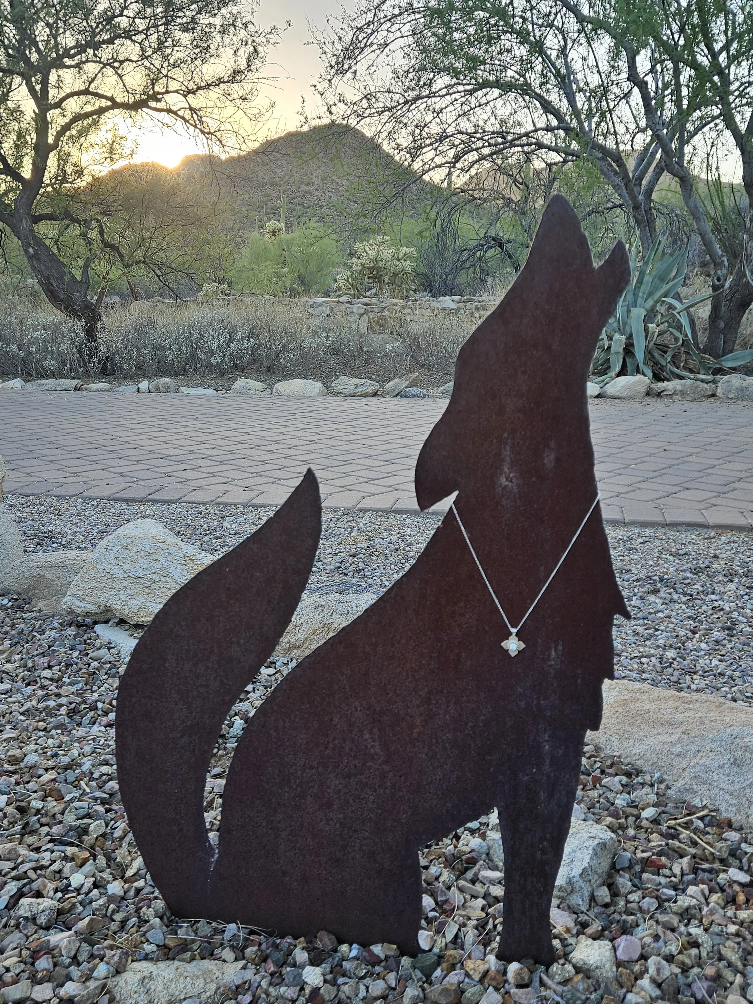 Rusty metal silhouette of a coyote wearing a diamond necklace in a desert landscape with trees, shrubs, rocks, and a mountain in the background, during sunset.