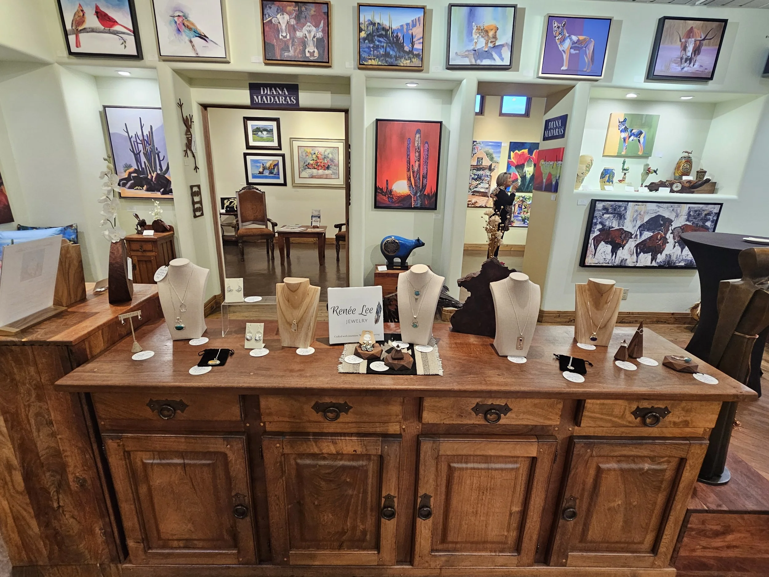 Interior view of an art gallery displaying paintings of animals, landscapes, and desert scenes, along with jewelry and decorative sculptures on a wooden table and shelves.