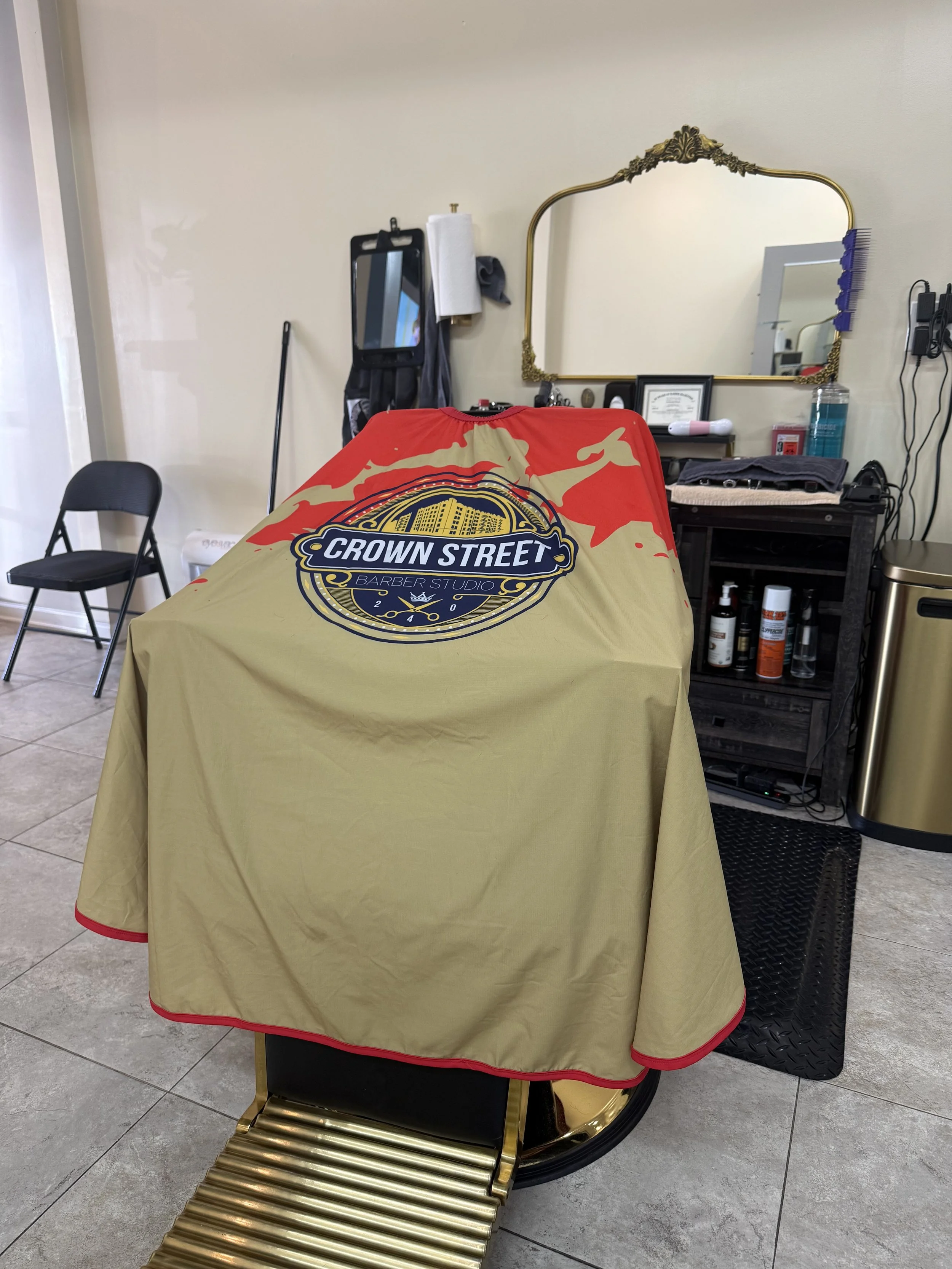 A barber shop chair covered with a cape that has 'Crown Street Barber Studio' logo. There's a black chair and barber tools in the background.