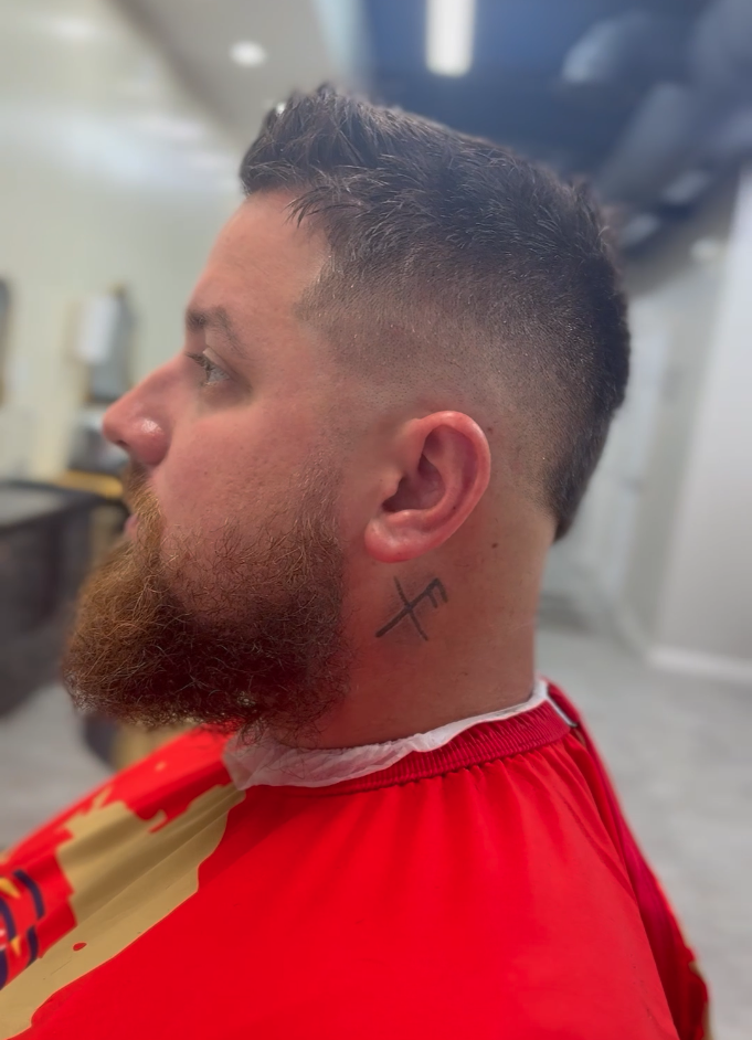 Man with short, fade hairstyle and beard, displaying a small cross tattoo on the side of his neck, wearing a red cape in a barbershop.