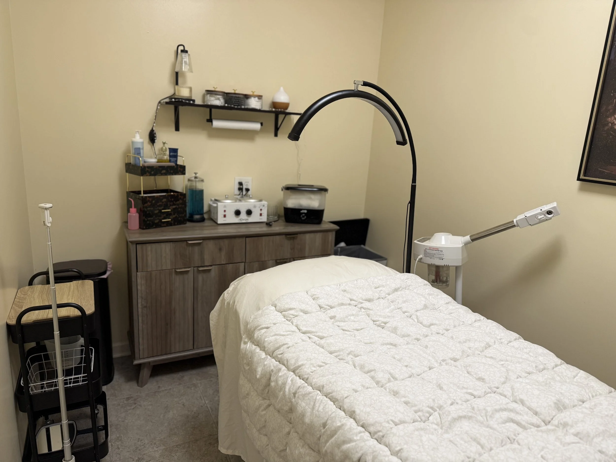 Medical examination room with a hospital bed covered with a white quilt, a wooden cabinet with medical supplies, a black lamp, and various medical equipment on the wall and countertops.