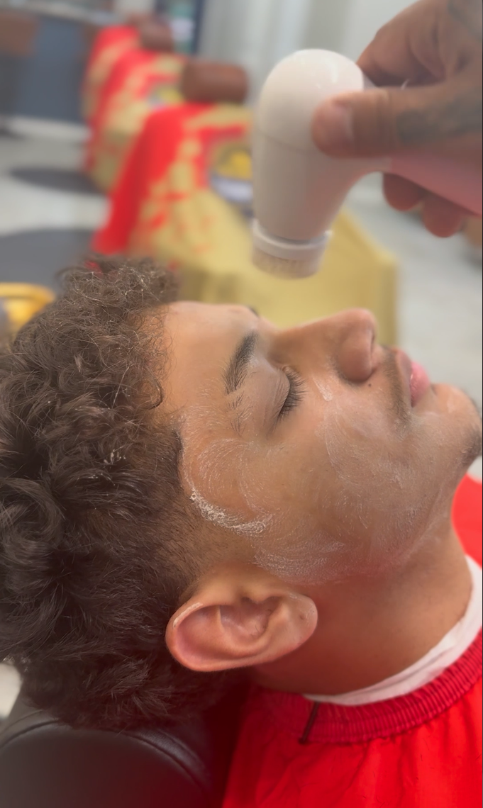 A man lying back with his eyes closed while a barber or stylist applies foam or shaving cream to his face. The stylist holds a white spray bottle or applicator above the man's face. The background shows blurred shelves or counters with barber supplie