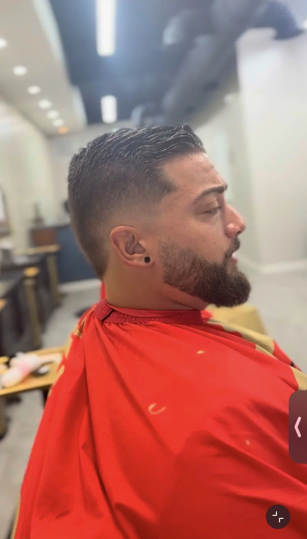 Man with short styled hair and beard sitting in a barbershop chair, wearing a red barber cape, with closed eyes.