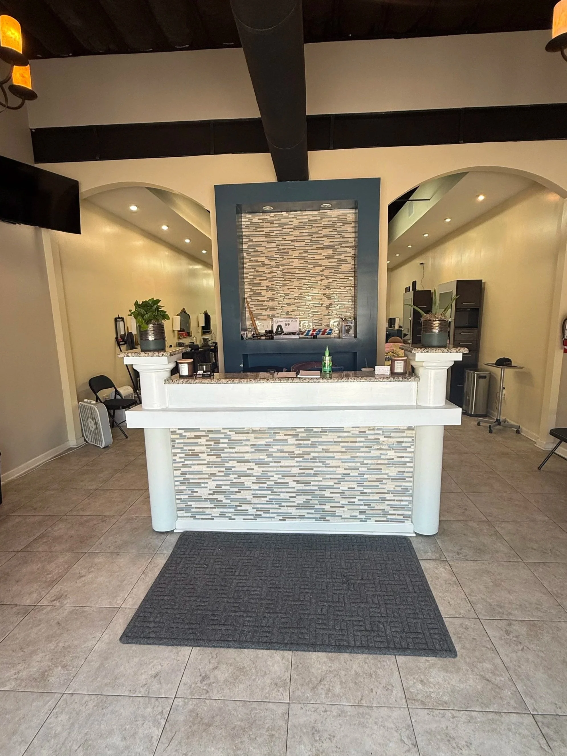 The image shows the front desk of a reception area with a tiled stone facade, potted plants, chairs, and a black rug in front of the desk. The background features a partial view of the reception space with equipment and furniture.
