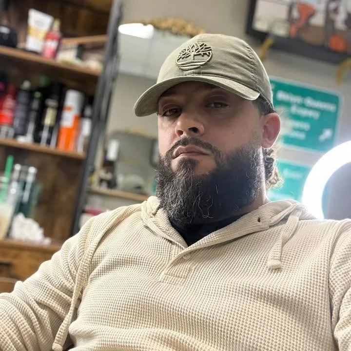 A man with a beard wearing a beige hoodie and a beige cap with a logo, sitting indoors with shelves and a bright light behind him.