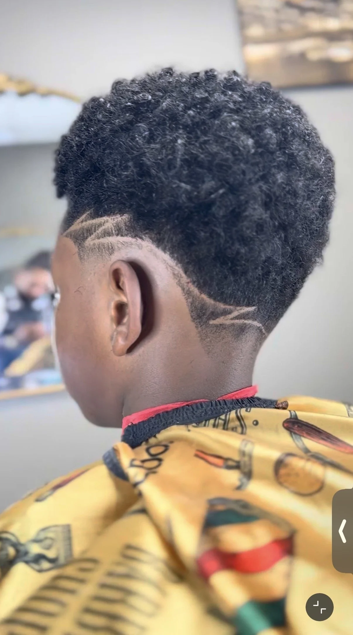 Back of a person's head with a stylish haircut featuring a shaved design and textured curly top, wearing a yellow barber cape with colorful patterns.