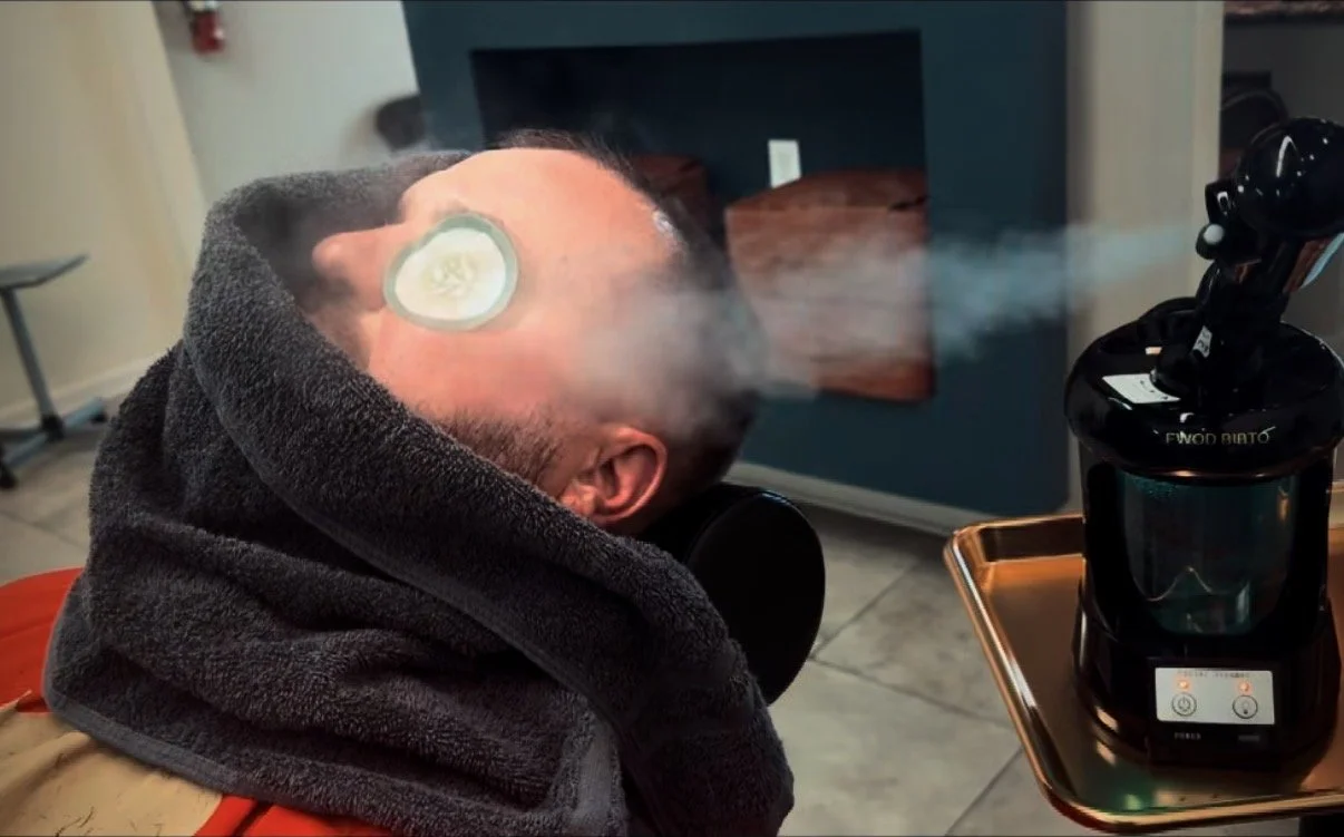 A man with a towel around his neck is using a vaporizer mask, exhaling vapor or steam in a room with a blue fireplace and a small table.