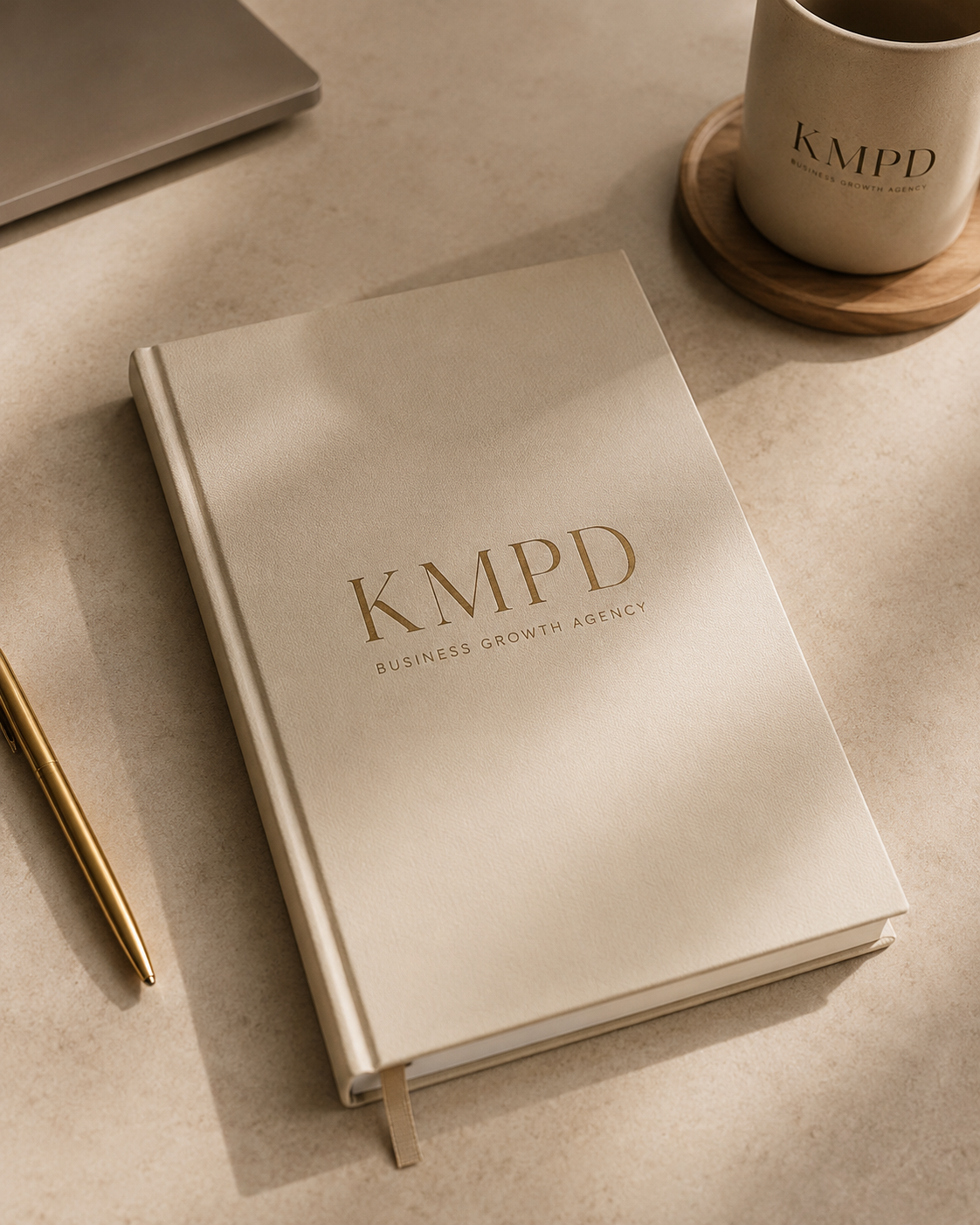 A beige notebook labeled 'KMPD Business Growth Agency' placed on a beige desk alongside a gold pen, a laptop, and a mug of coffee on a wooden coaster.