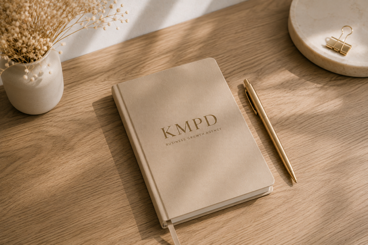 A beige notebook labeled 'KMPD Business Growth Agency,' a gold pen, a marble tray with a gold binder clip, a white vase with dried flowers on a wooden desk.