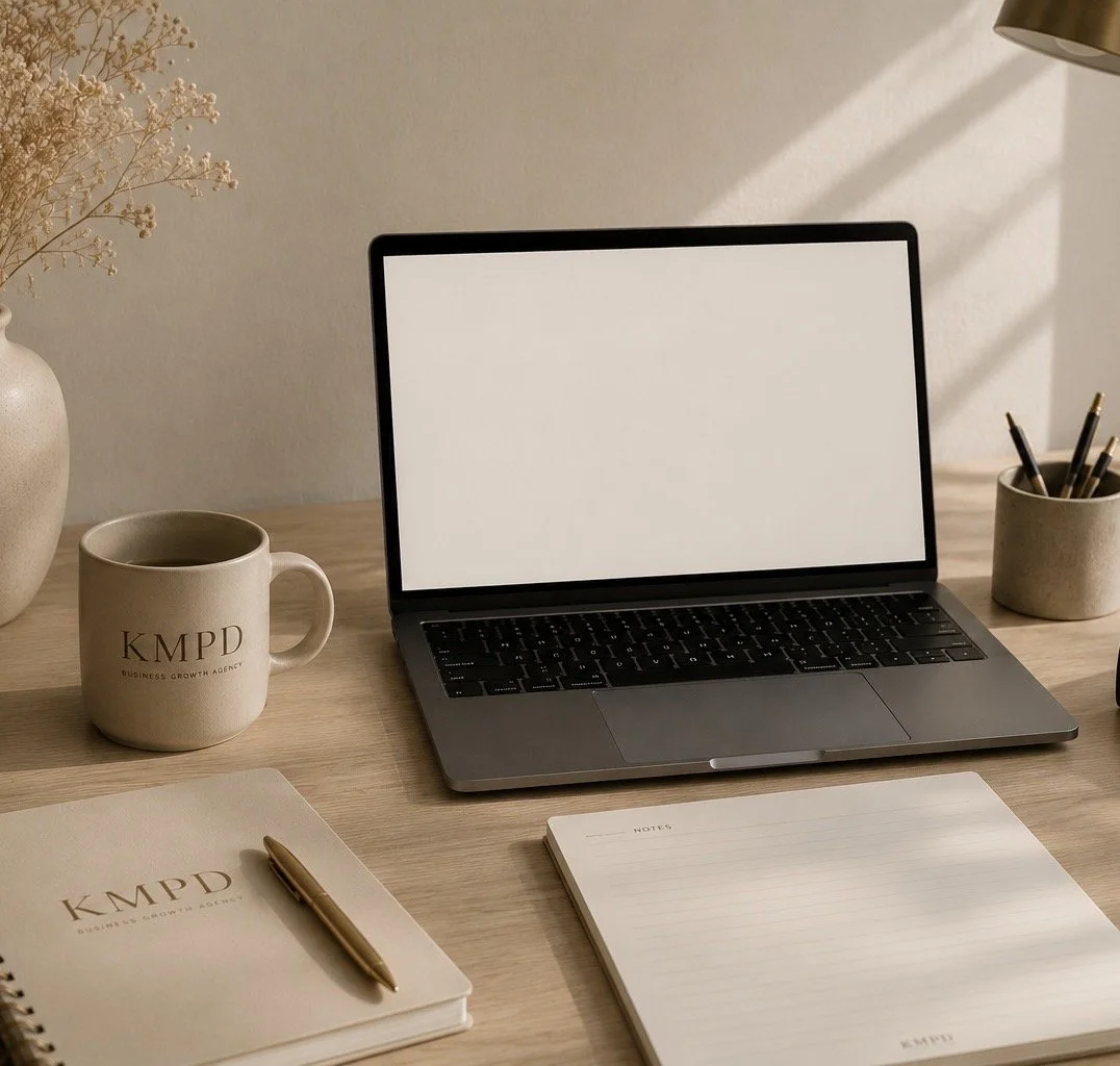 A neatly organized workspace with a laptop that has a blank white screen, a coffee mug labeled 'KMPD Business Growth Agency', a notebook and a pen, a notepad, a cup with pens, and a vase of dried flowers on a wooden desk.