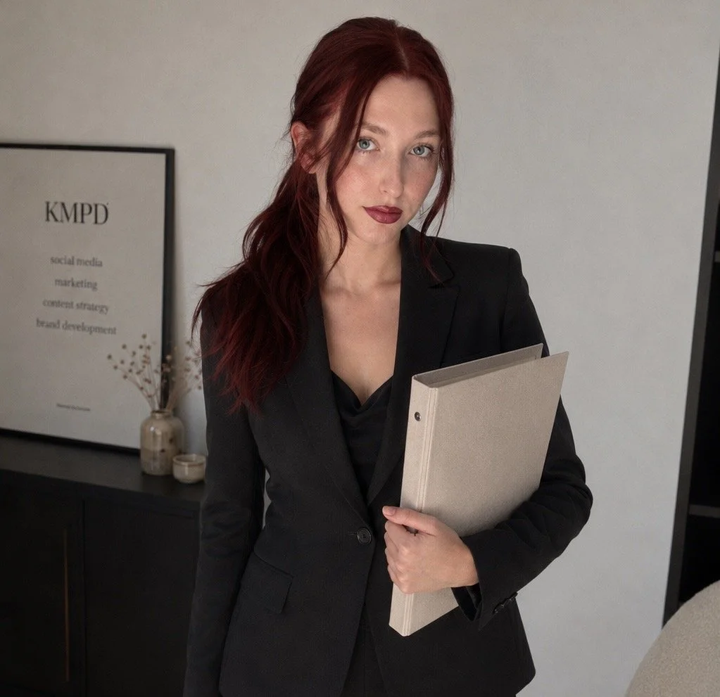 A woman with long red hair and wearing a black blazer, holding a beige folder, standing in an office with a sign that reads 'KMPD' in the background.