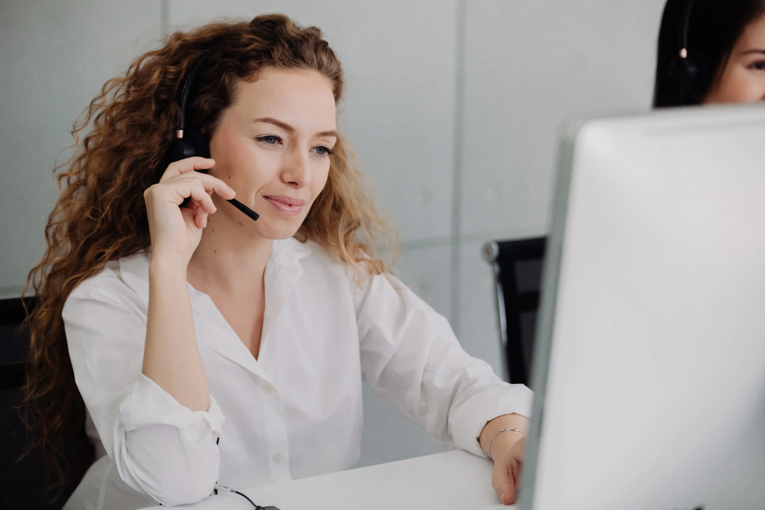A woman with curly red hair wearing a white blouse and headset, working at a computer in an office setting.