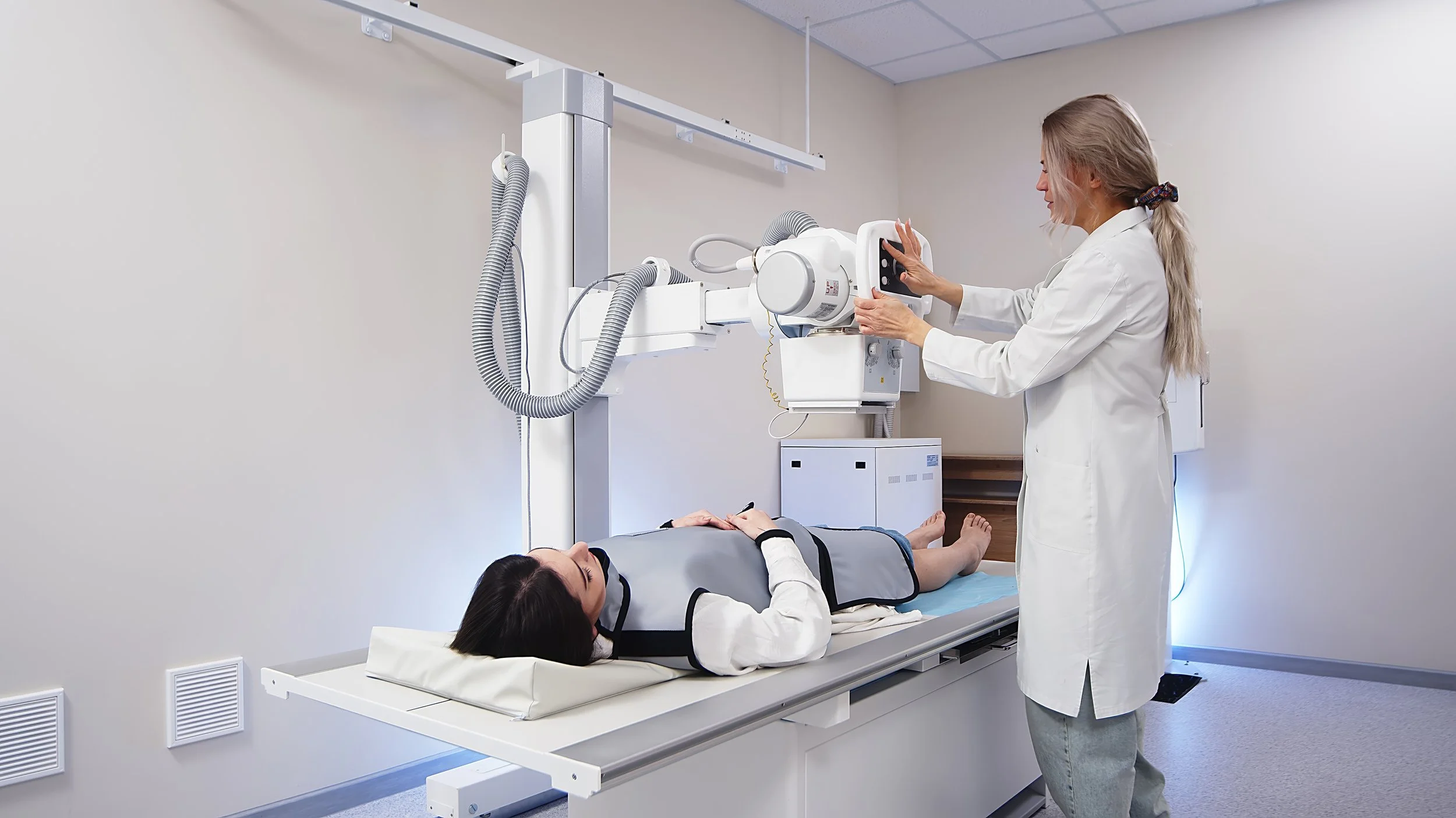 Medical technician adjusting X-ray machine over patient lying on table in hospital room.