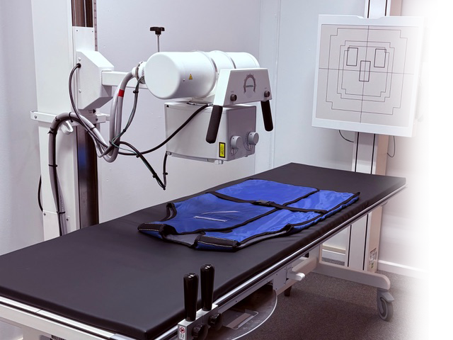 Medical imaging room with X-ray machine and examination table covered with blue lead apron.