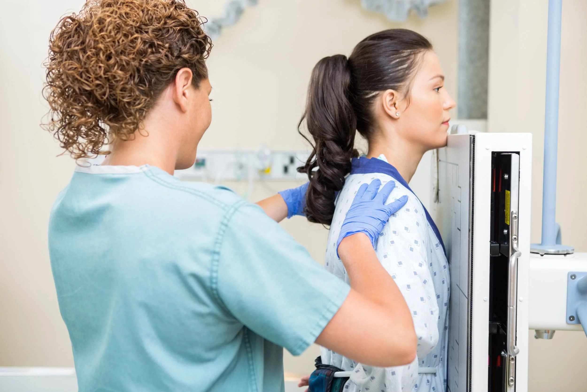 A medical professional is assisting a woman who is standing, possibly for a breast imaging or mammogram procedure, in a medical setting.