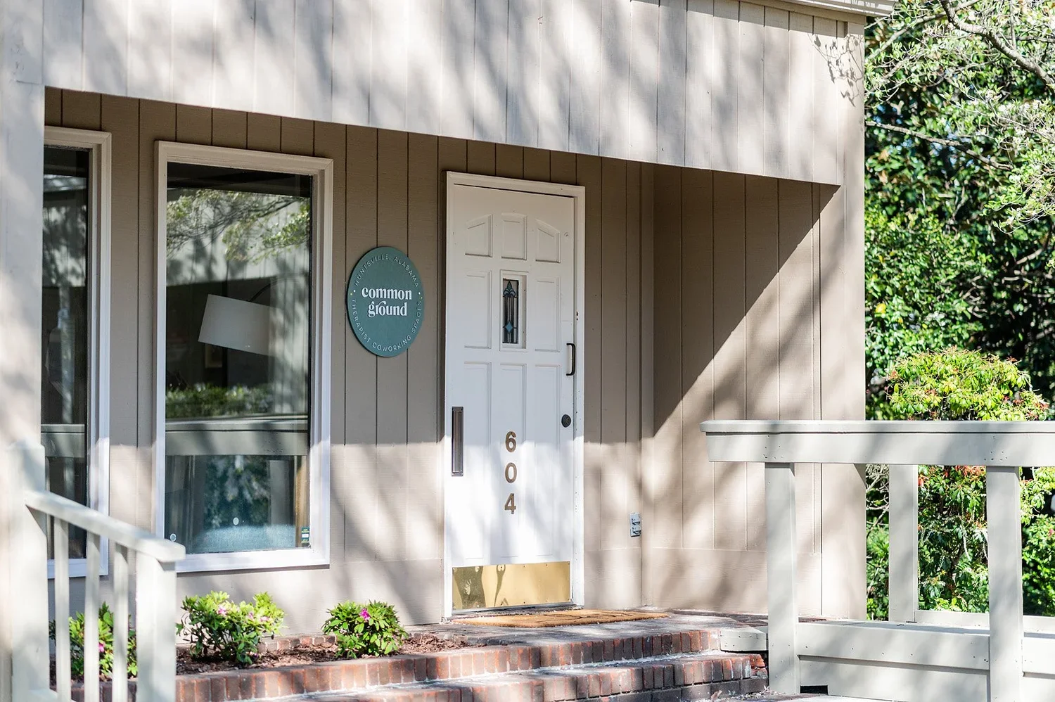 Front door of a building with the number 604, a small window in the door, and a sign that reads "common ground" with additional text about coworking spaces. The building has beige vertical siding and nearby greenery.