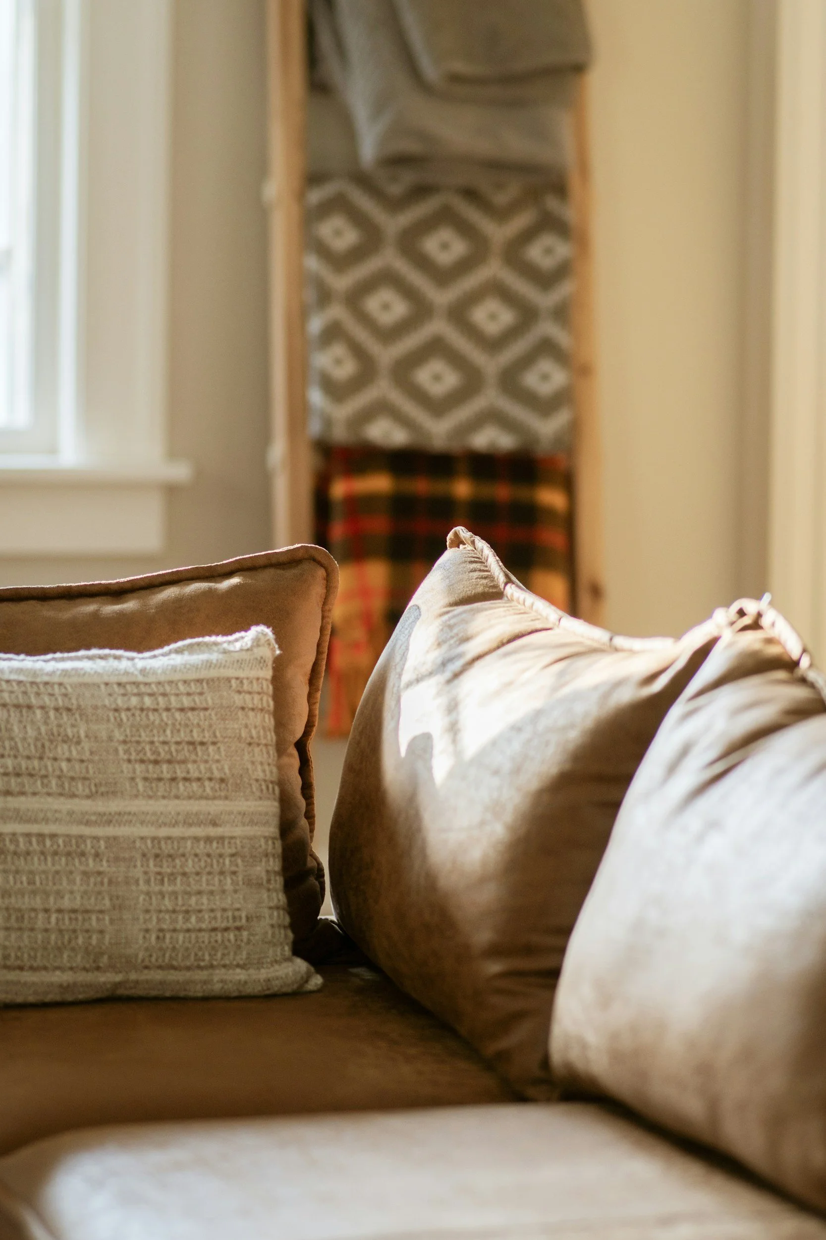 Close-up of a row of pillows on a couch in a cozy living room, with a window nearby and a decorative blanket hanging behind, illuminated by natural light.