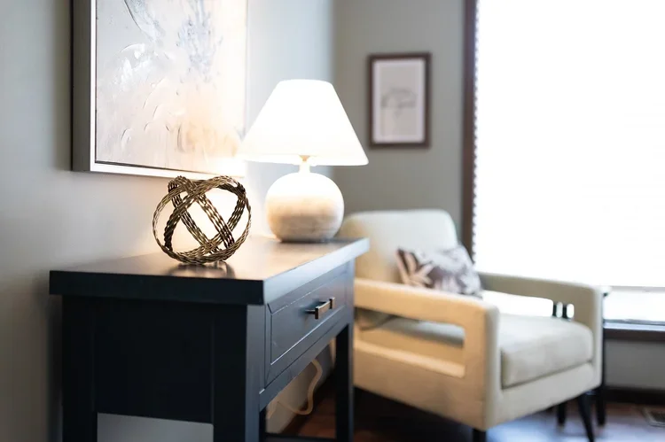 A therapy room corner with a black side table, a white table lamp, a decorative wicker sphere, a white armchair with a patterned pillow, and a window with blinds.