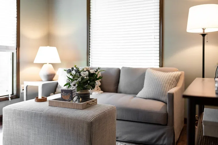 A therapy room with a gray sofa, striped pillow, coffee table with a flower arrangement, side table with a lamp, and blinds on the window.