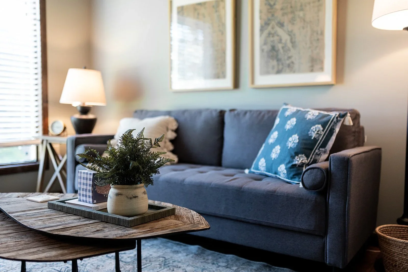 A therapy room with a grey sofa, pillows, a wooden coffee table with a plant in a white vase, and a side table with a lamp and a clock. Two framed artworks hang on the wall, and a window with wooden blinds is visible.