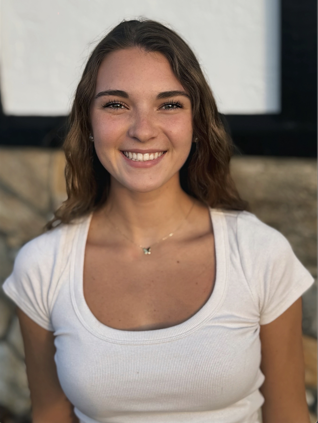 Close-up of a young woman smiling, with light brown, wavy hair, wearing a white t-shirt and butterfly necklace, in front of a blurred background.