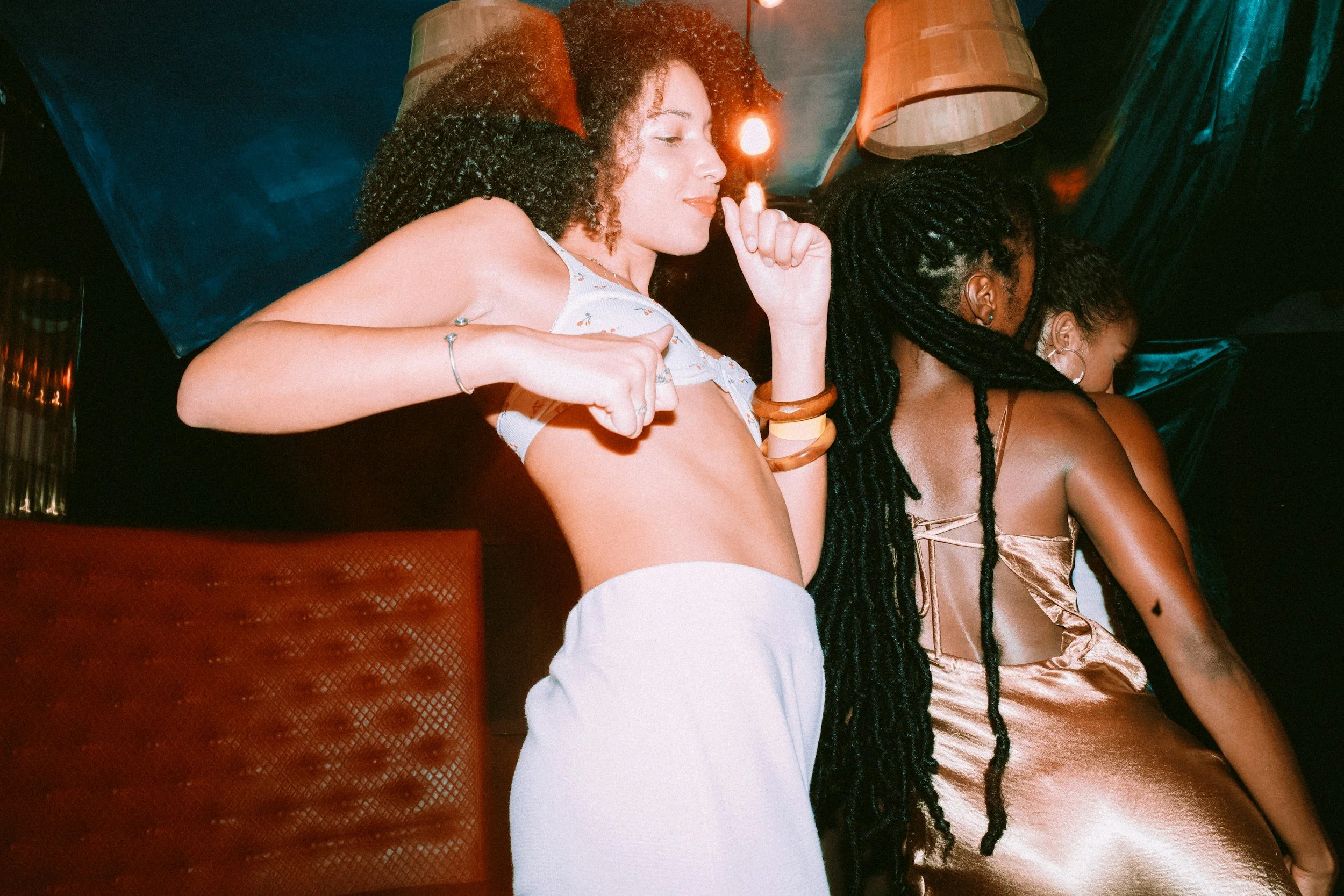 Two women at a party or nightclub with one woman in a floral top and the other in a satin dress, both with curly and braided hair, enjoying a moment together.