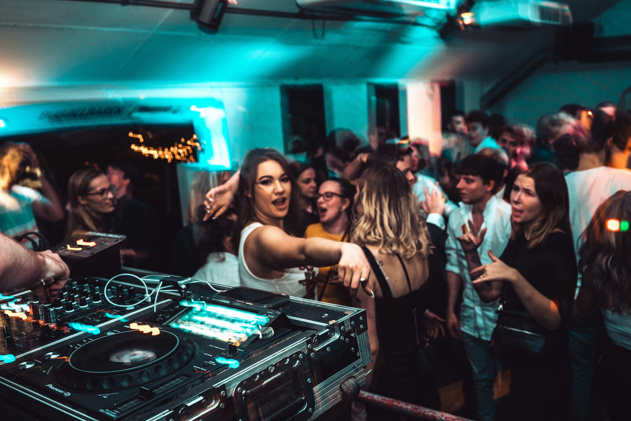 A crowded dance party with a DJ mixing music on a turntable, and people dancing and socializing in a dimly lit room with blue and pink lighting.
