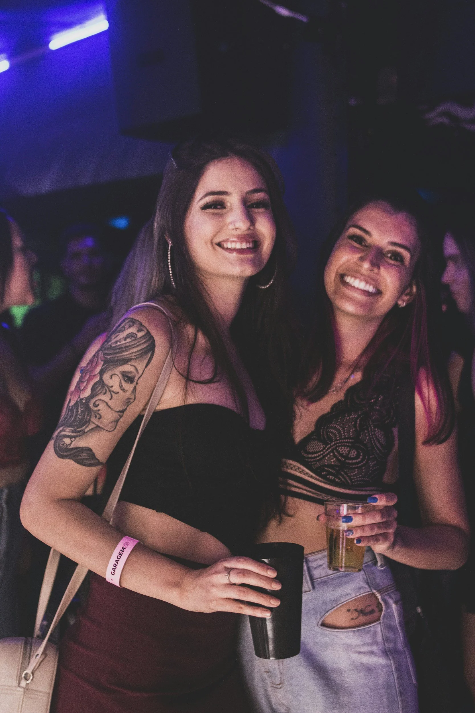Two women smiling at a party with colorful lights. One woman has tattoos on her arm, long dark hair, and is holding a drink. The other woman has curly dark hair, is wearing a black lacy top, and is also holding a drink.