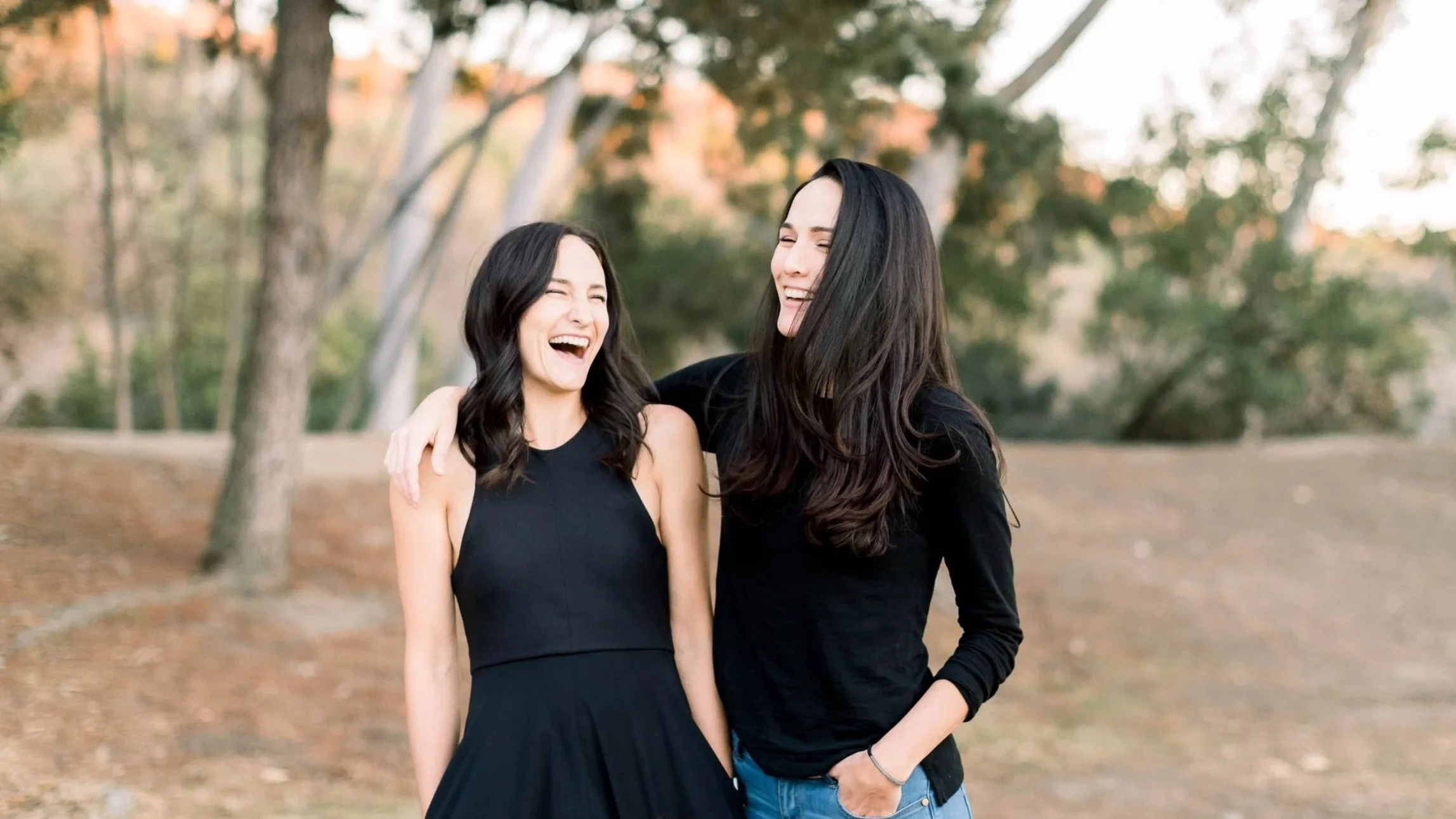Two women laughing and enjoying each other's company outdoors in a park, with trees and autumn leaves in the background.