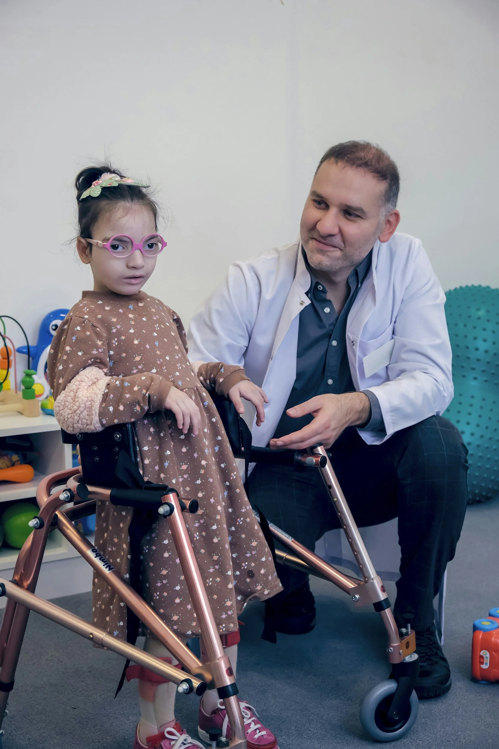 A young girl in a brown dress with floral patterns and pink sneakers in a wheelchair, talking with a male doctor in a medical office. The girl is wearing pink glasses and a green headband, and there are colorful toys in the background.