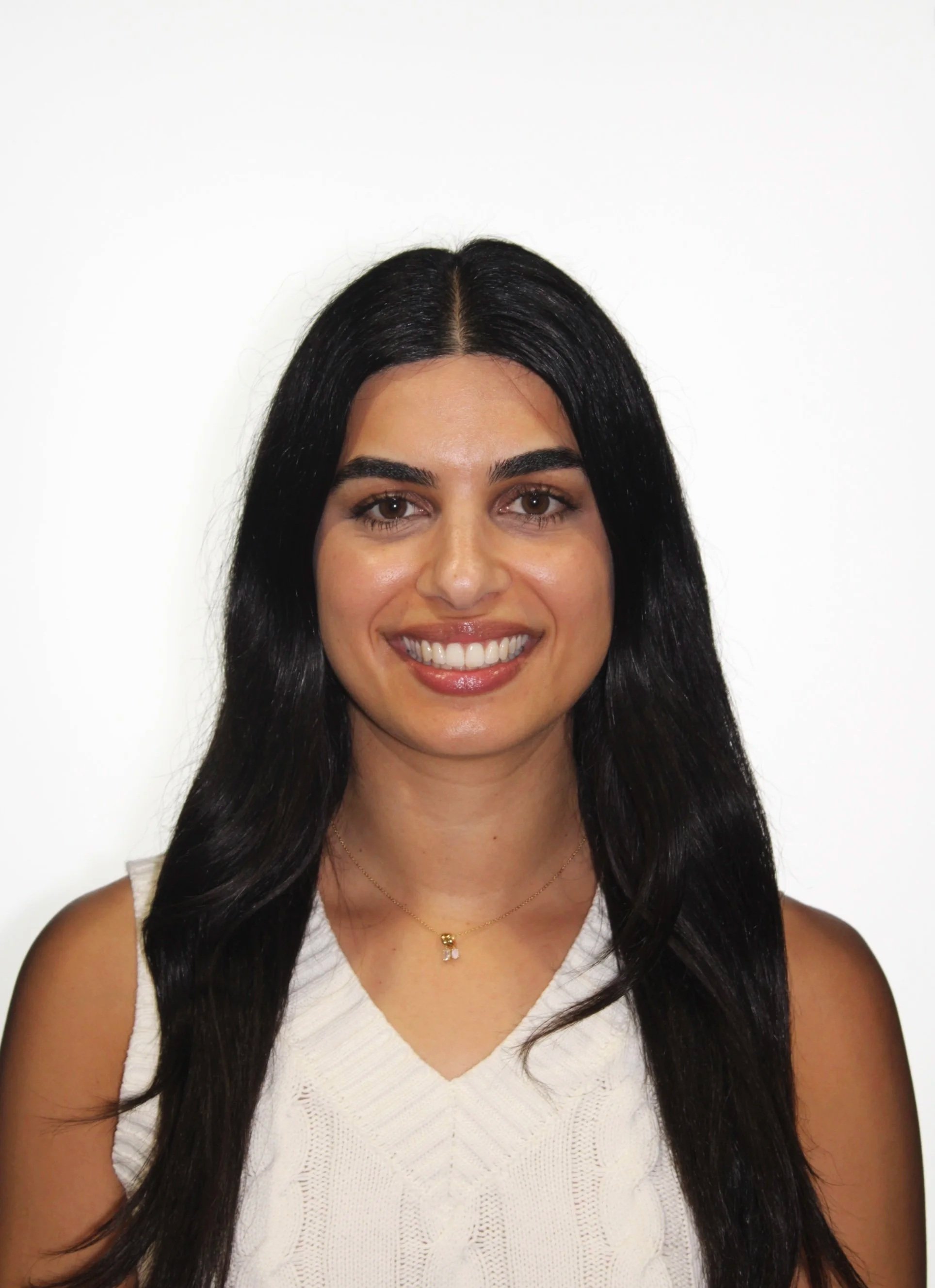 A woman with long black hair smiling, wearing a white sleeveless top and a gold necklace, standing against a white background.
